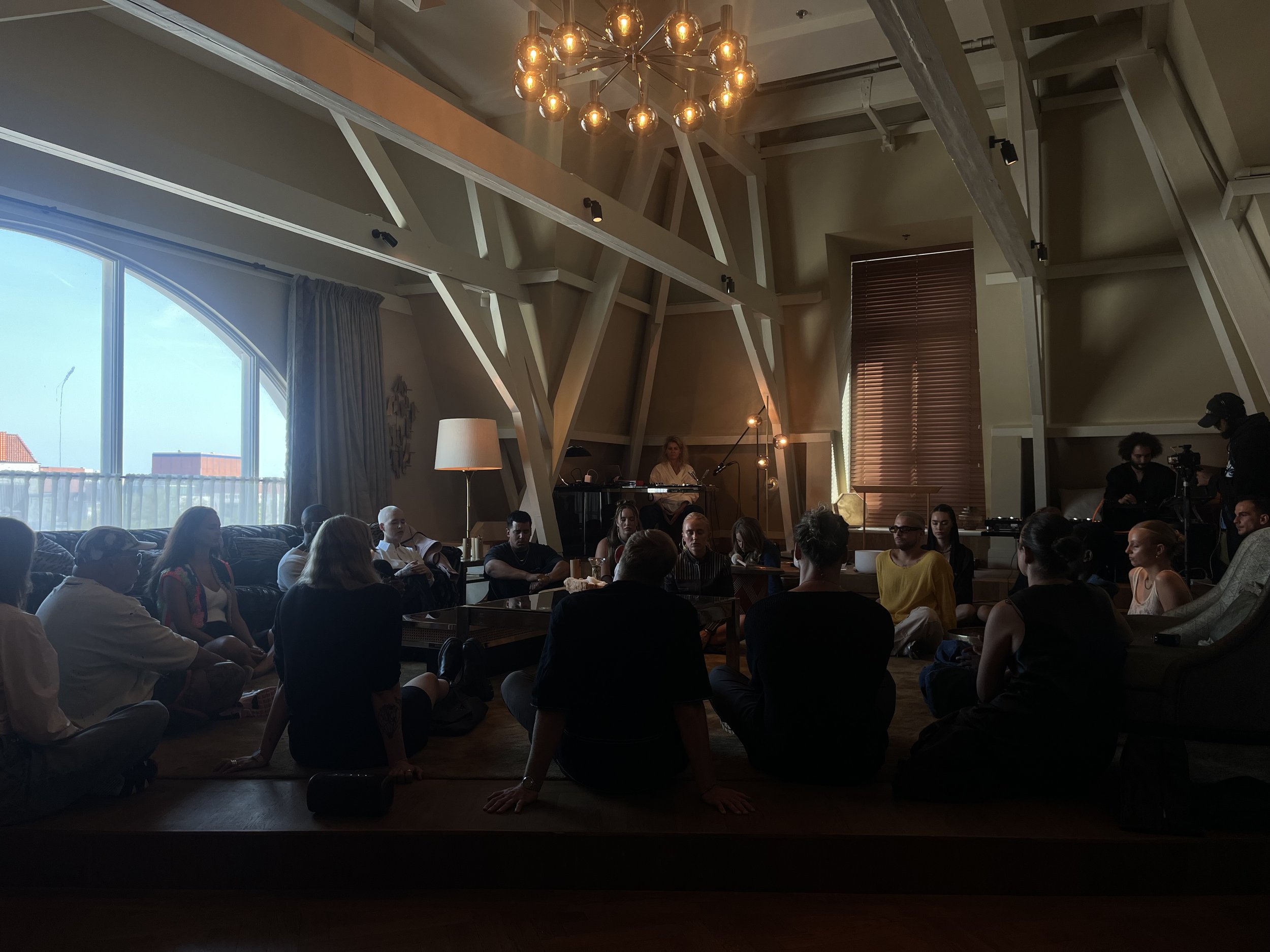 People sitting on the floor in a large room with wooden floors, a large window, and a modern chandelier, attending an event or gathering.