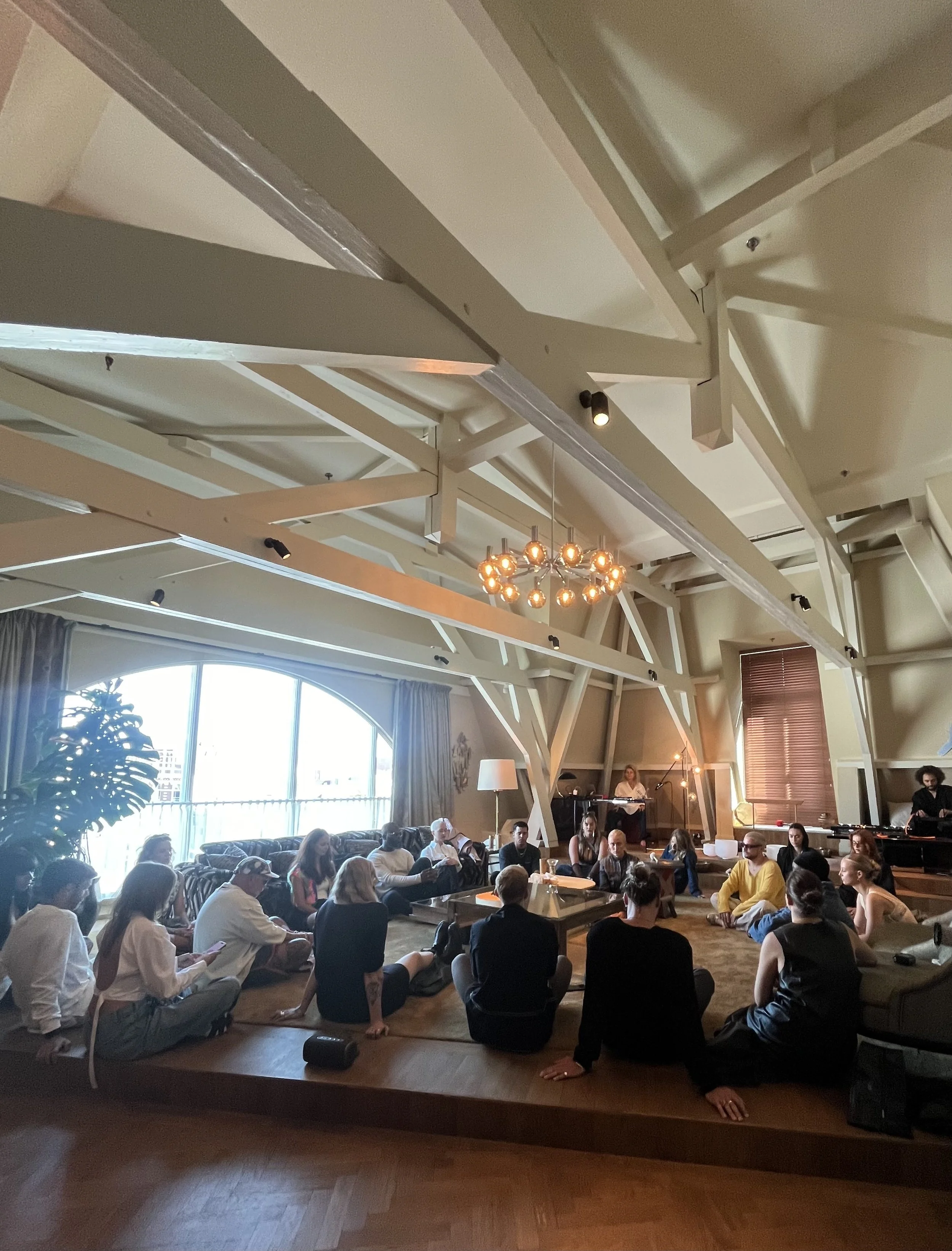 People attending a seated event in a spacious room with large arched windows, white ceiling beams, and a central chandelier with exposed bulbs, while some sit on the floor.