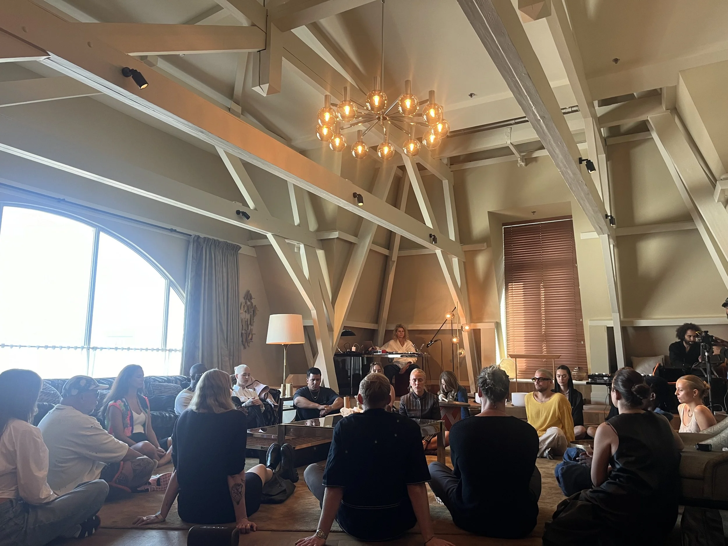 A group of people gathered in a cozy, modern room with a high ceiling, large window, and warm lighting. They are sitting and standing, listening to a woman at a piano, with some sitting on the floor and others on chairs. The room features wooden beams, a chandelier, and modern decor.