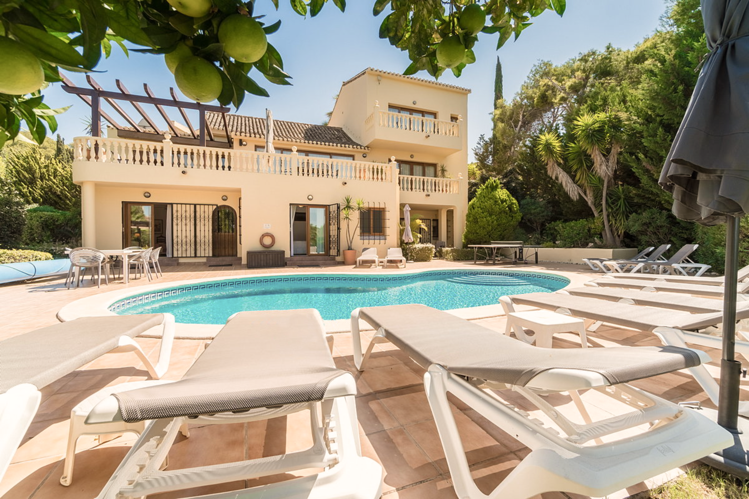 Residential house with a balcony, a swimming pool, and poolside lounge chairs surrounded by greenery and trees.