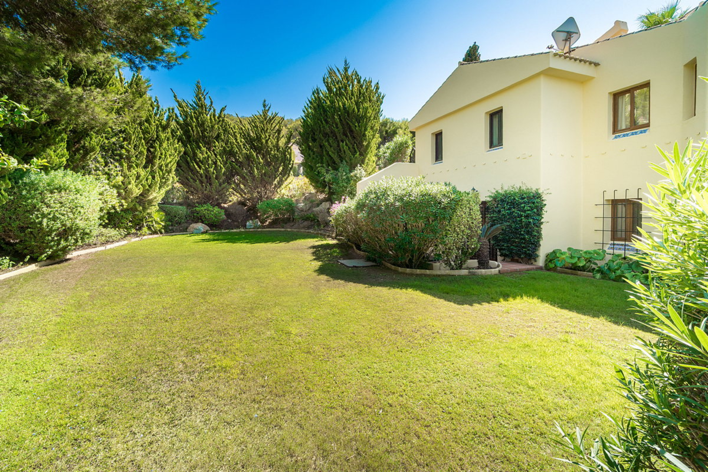 A backyard with a well-maintained grassy lawn, various green bushes, and tall trees surrounding a light-colored house with windows, a satellite dish on the roof, and a clear blue sky.