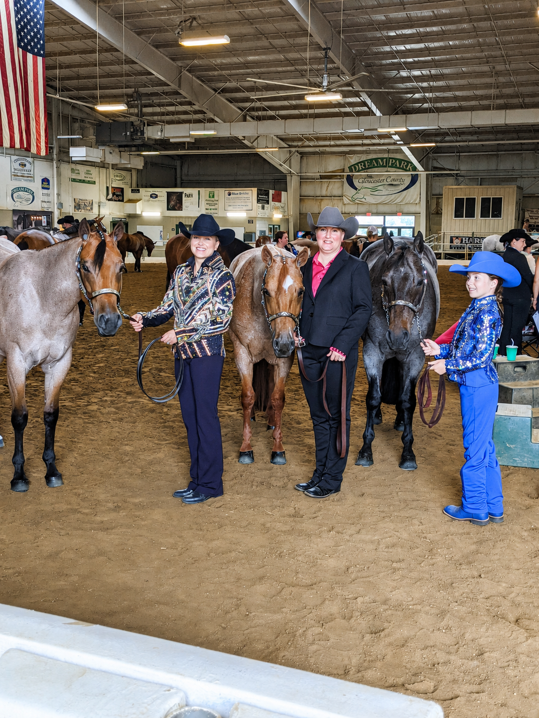 Three women and a girl standing with horses inside an indoor arena at a rodeo or horse show, with American flag and various signs in the background.