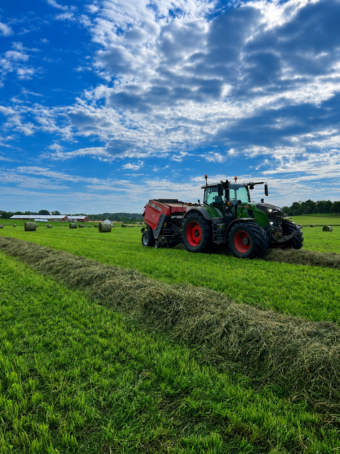 A tractor working in a green field with hay bales, under a blue sky with scattered clouds.