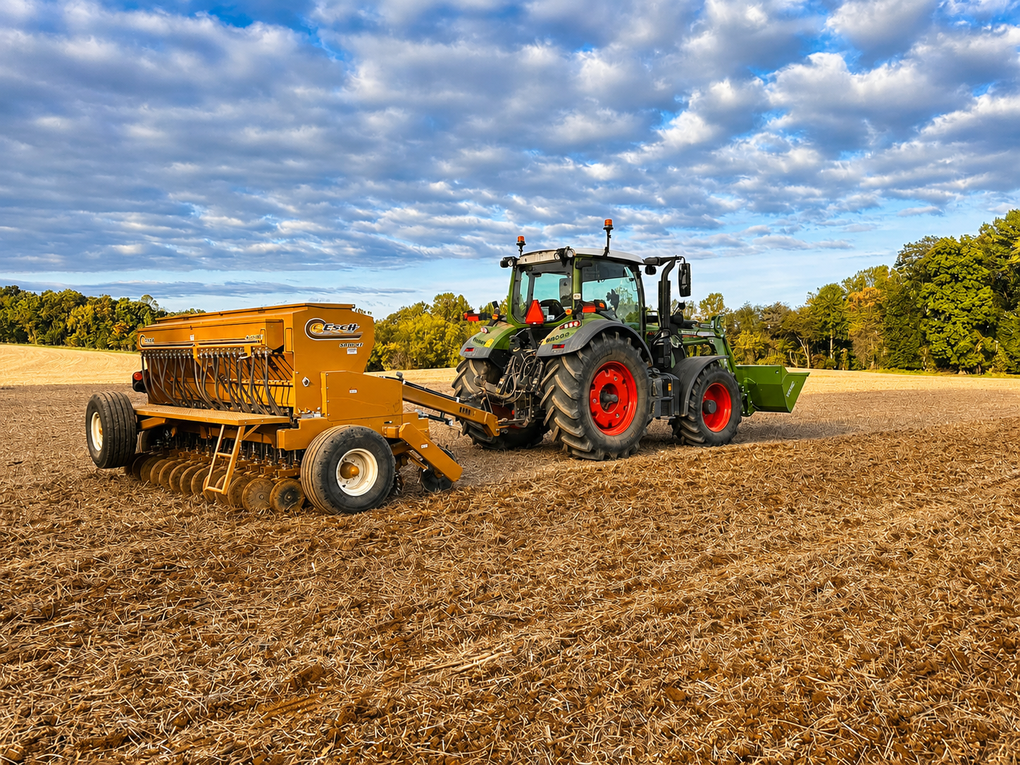 A tractor on a farm field with a seed planter attached, under a partly cloudy sky, surrounded by trees.