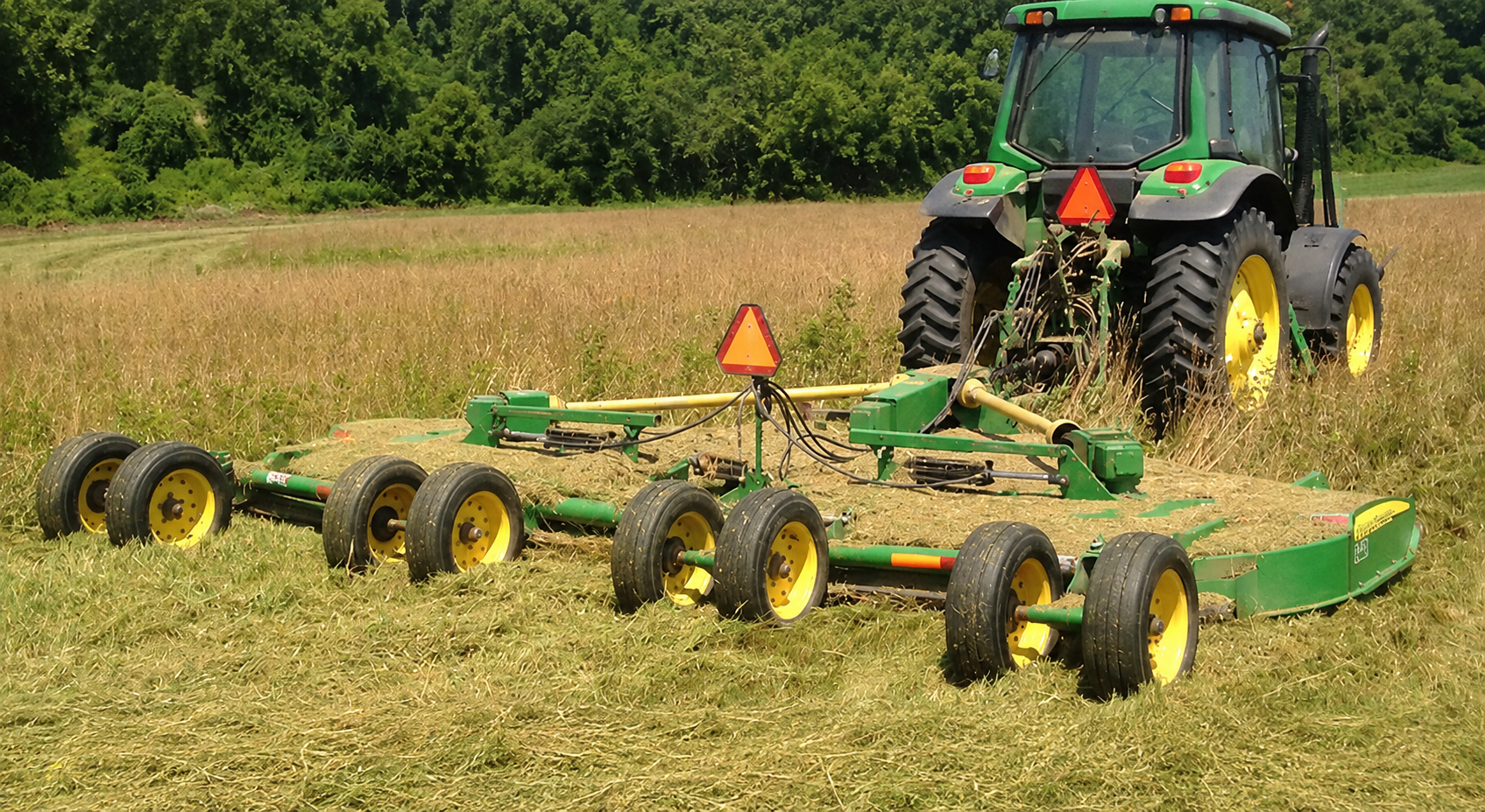 A green tractor towing a green and yellow agricultural implement in a field, with trees in the background.
