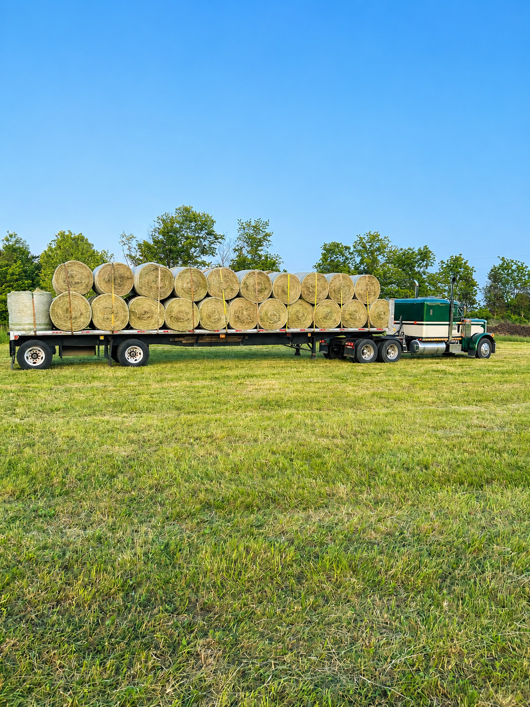 A flatbed trailer attached to a semi-truck sits in a grassy field under a clear blue sky, carrying large rolled hay bales.