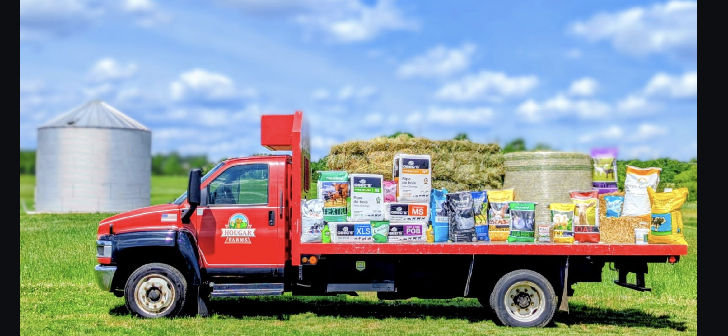 Red farm truck carrying bags of animal feed on a green field, with a silo and hay bales in the background under a blue sky with clouds.