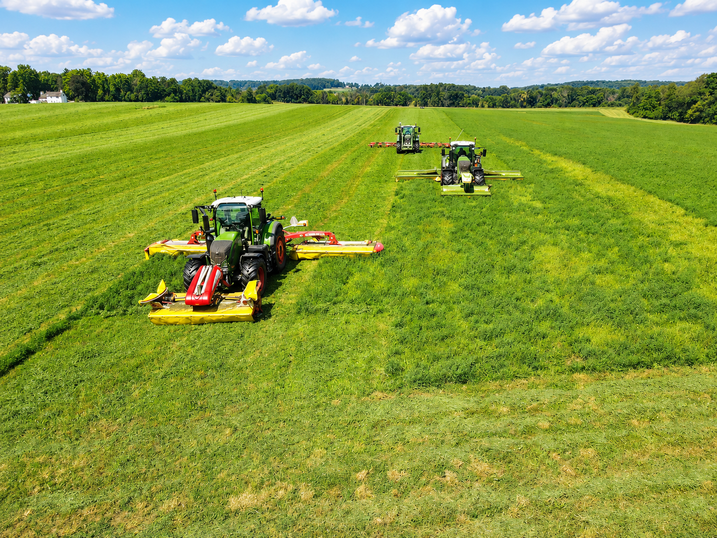 Three tractors are mowing a large green field of grass under a bright blue sky with scattered clouds.