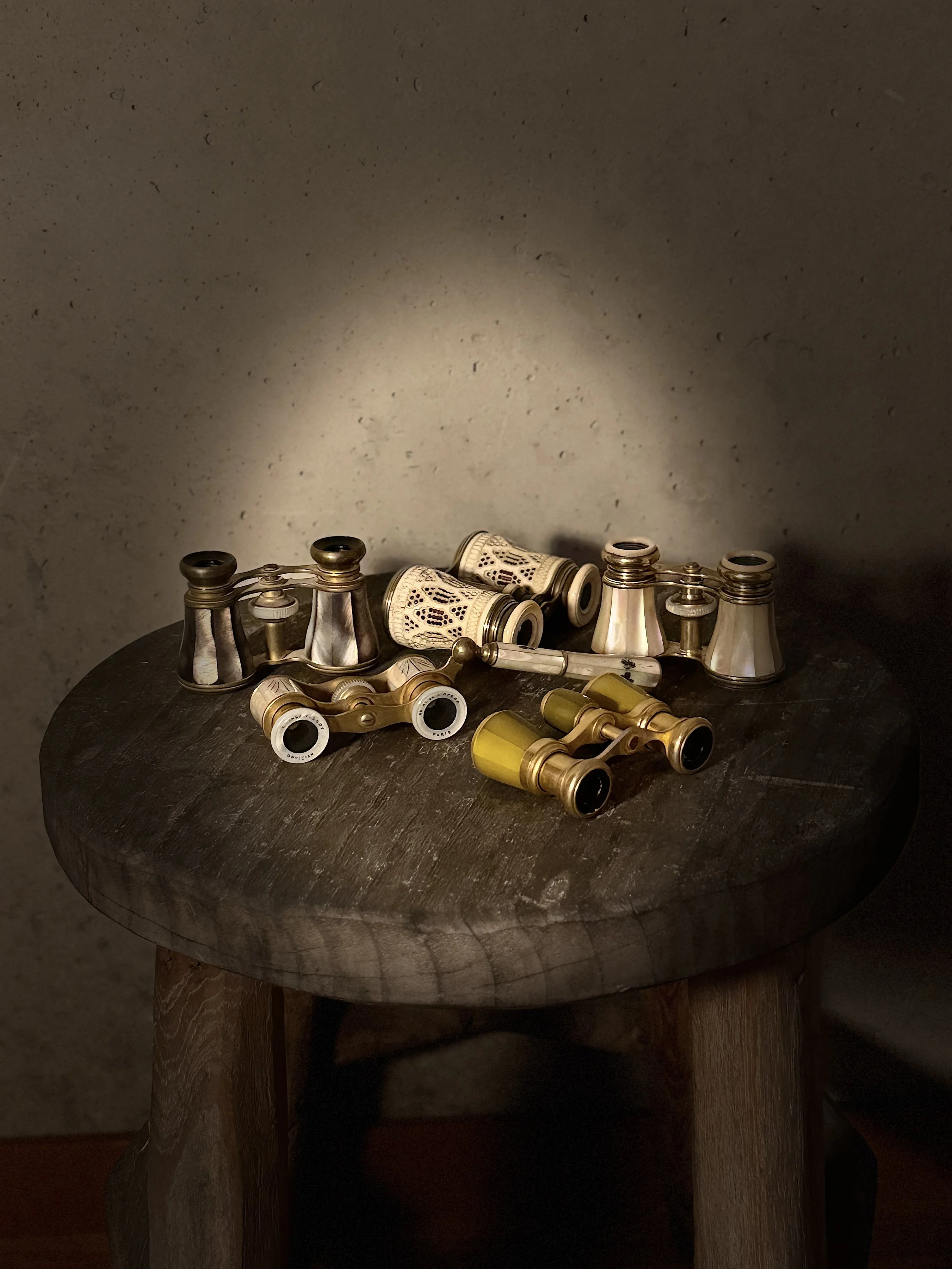 Assorted antique binoculars and a monocular placed on a dark wooden round table, with a plain wall in the background.
