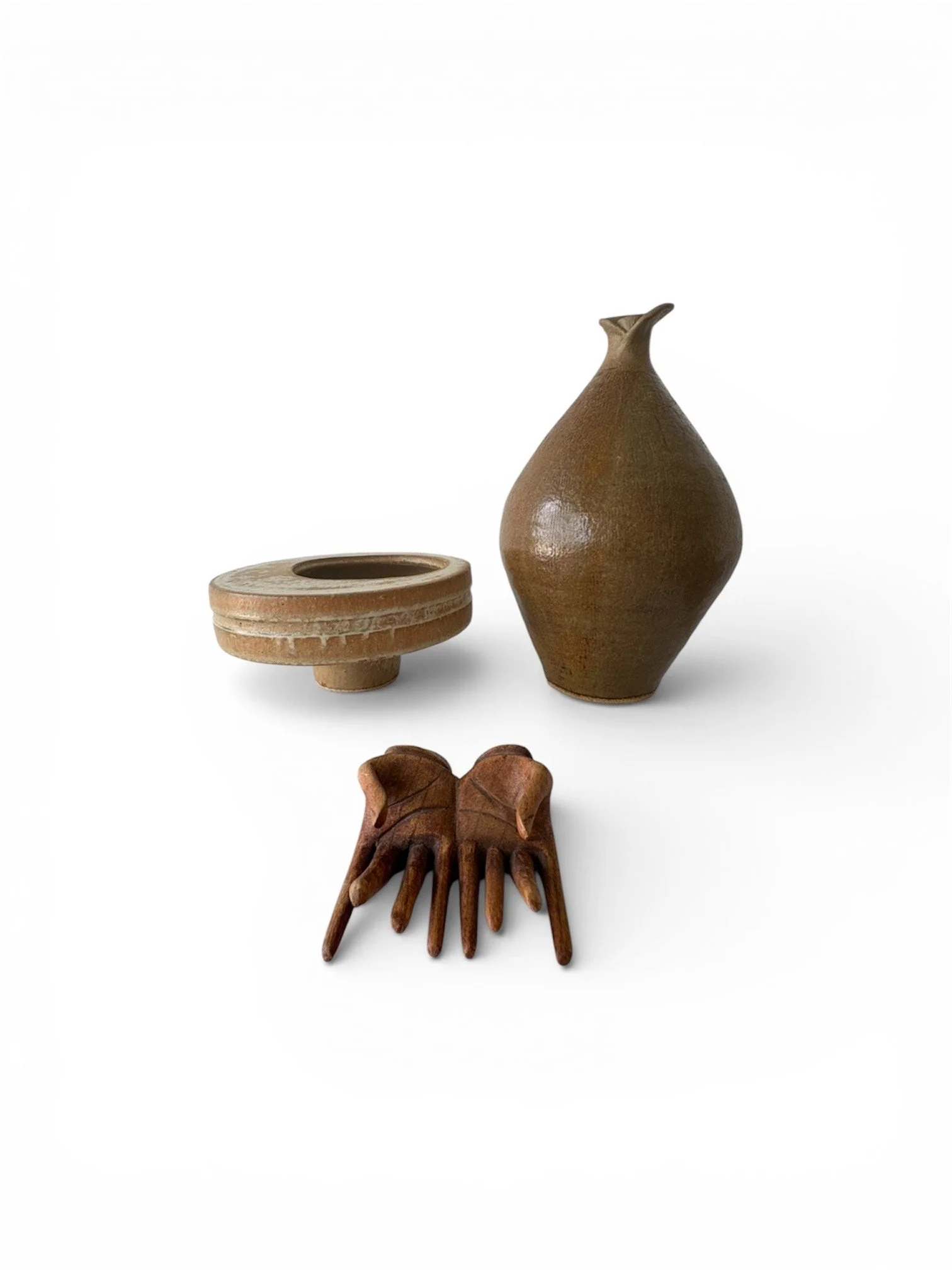 Three decorative objects including a carved hand-shaped object, a small disk-shaped ikebana potteryvessel, and a tall, curved studio pottery vessel with a small spout on top, arranged on a white background.