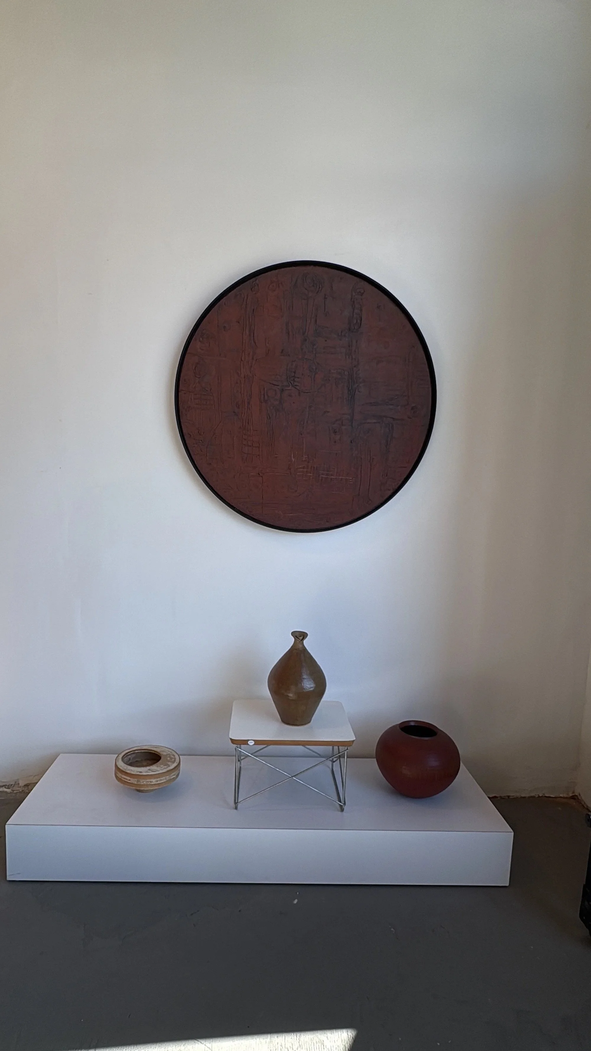 Display of various pottery vases and bowls on a white table, with a large round dark artwork hanging on the wall behind them.
