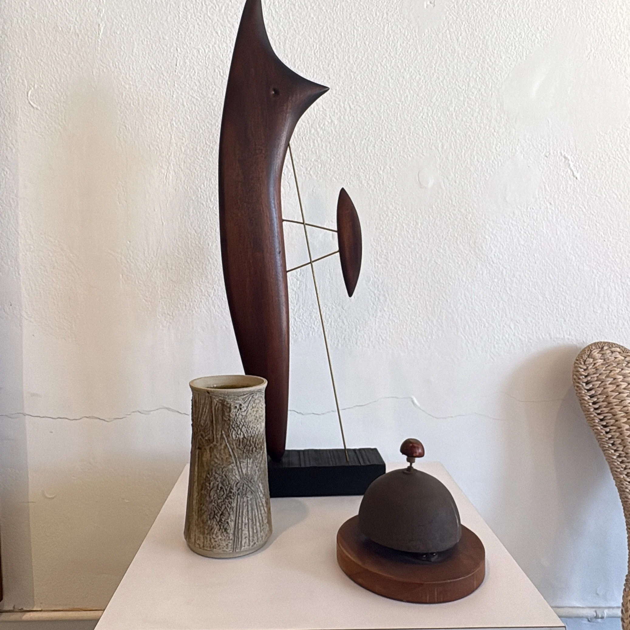 A collection of objects including a large wooden mid century modernist  sculpture, a studio pottery cessel with intricate carvings, a  dark wooden base bell,  on a white surface.