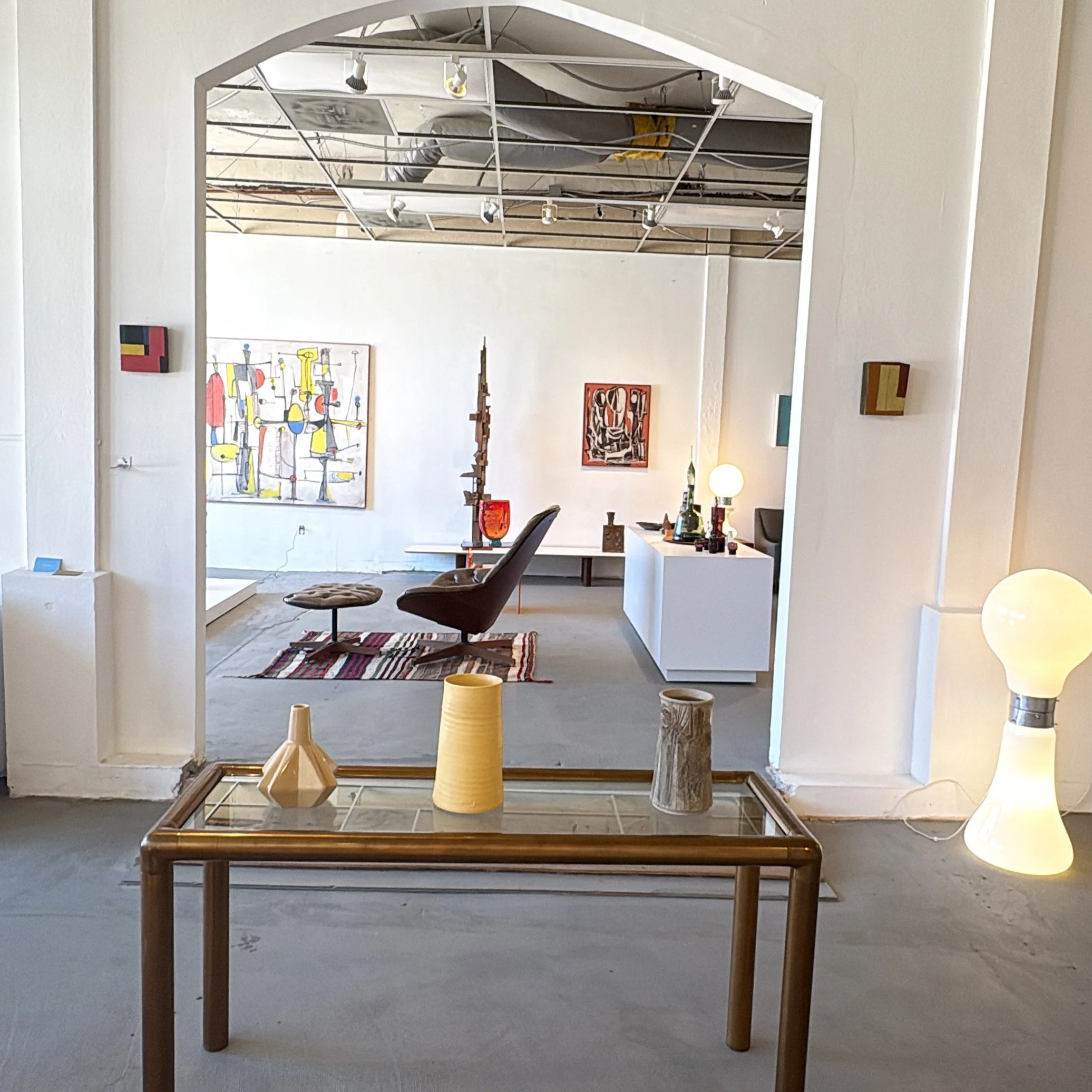 Interior view of an art gallery showing various paintings on the walls and sculptures on display, with a large mirror reflecting the ceiling.