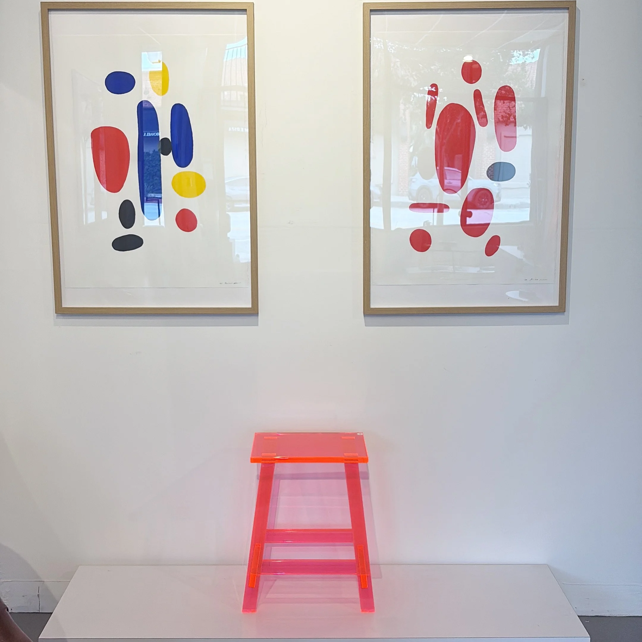 Contemporary art installation featuring three vintage gouache paintings  with abstract shapes in primary colors, over a white background, framed in wood. A pink transparent lucite stool is positioned in front of the artwork.