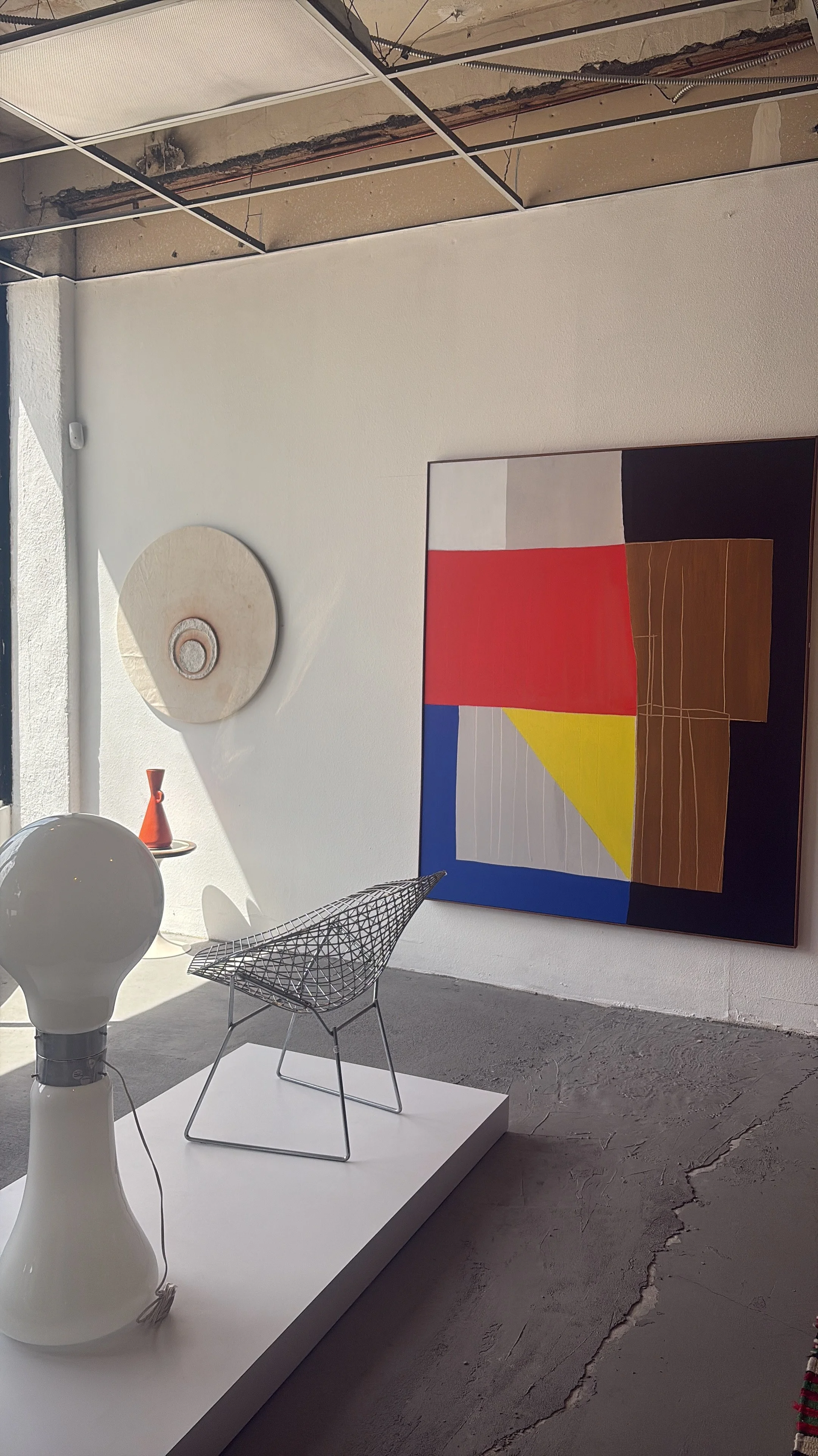 An art gallery display featuring a geometric abstract painting with blocks of red, yellow, blue, black, gray, and white. The display area includes a white platform with a white lamp, a wire chair, and a ceramic vase. The gallery space has an unfinished ceiling and a textured concrete floor.