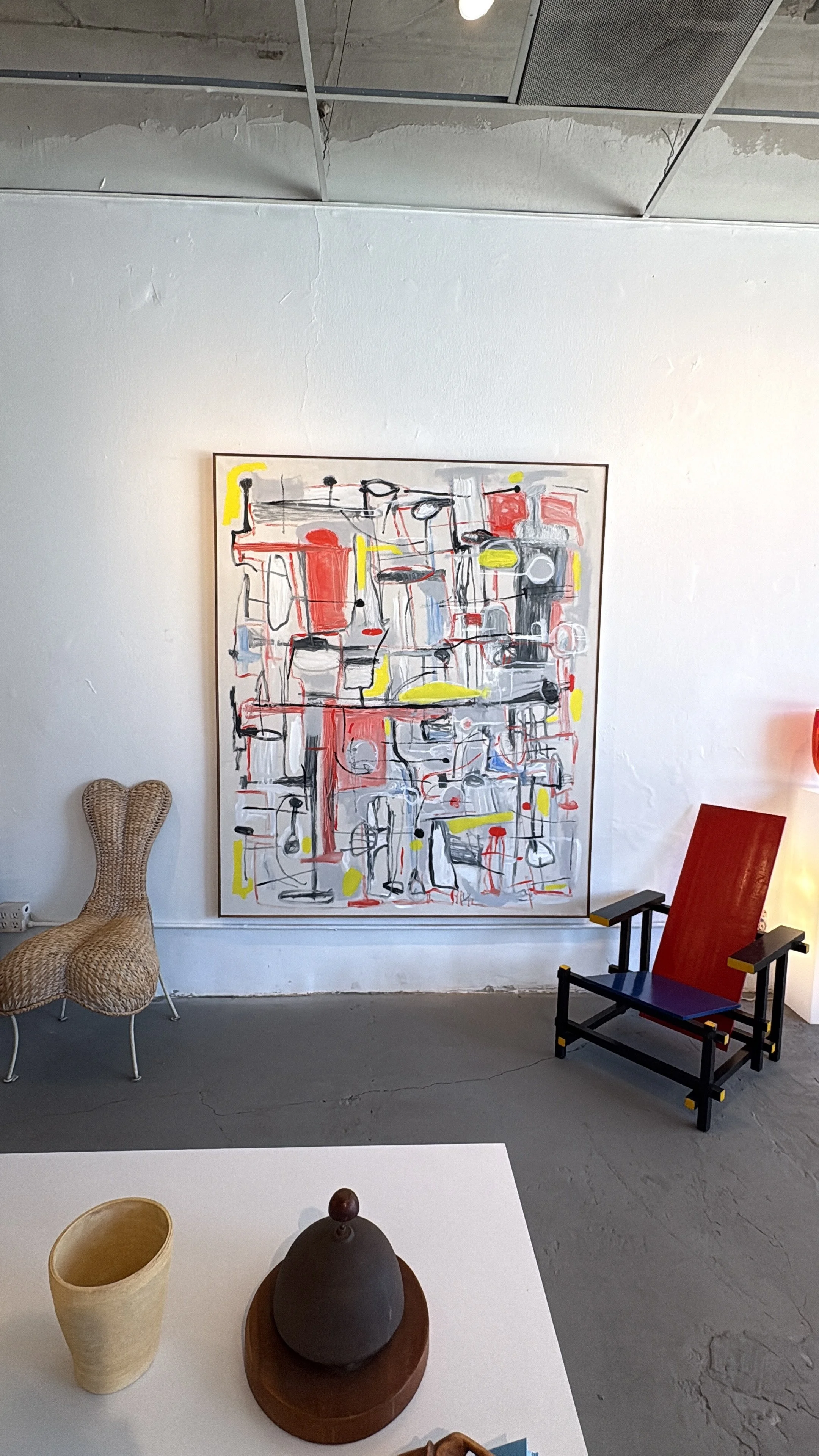 A white table with vintage pieces of pottery and an antique service bell on display plinth in the foreground. In the background, there is a colorful abstract painting on the wall, a wicker chair to the left, and a red and black chair to the right.