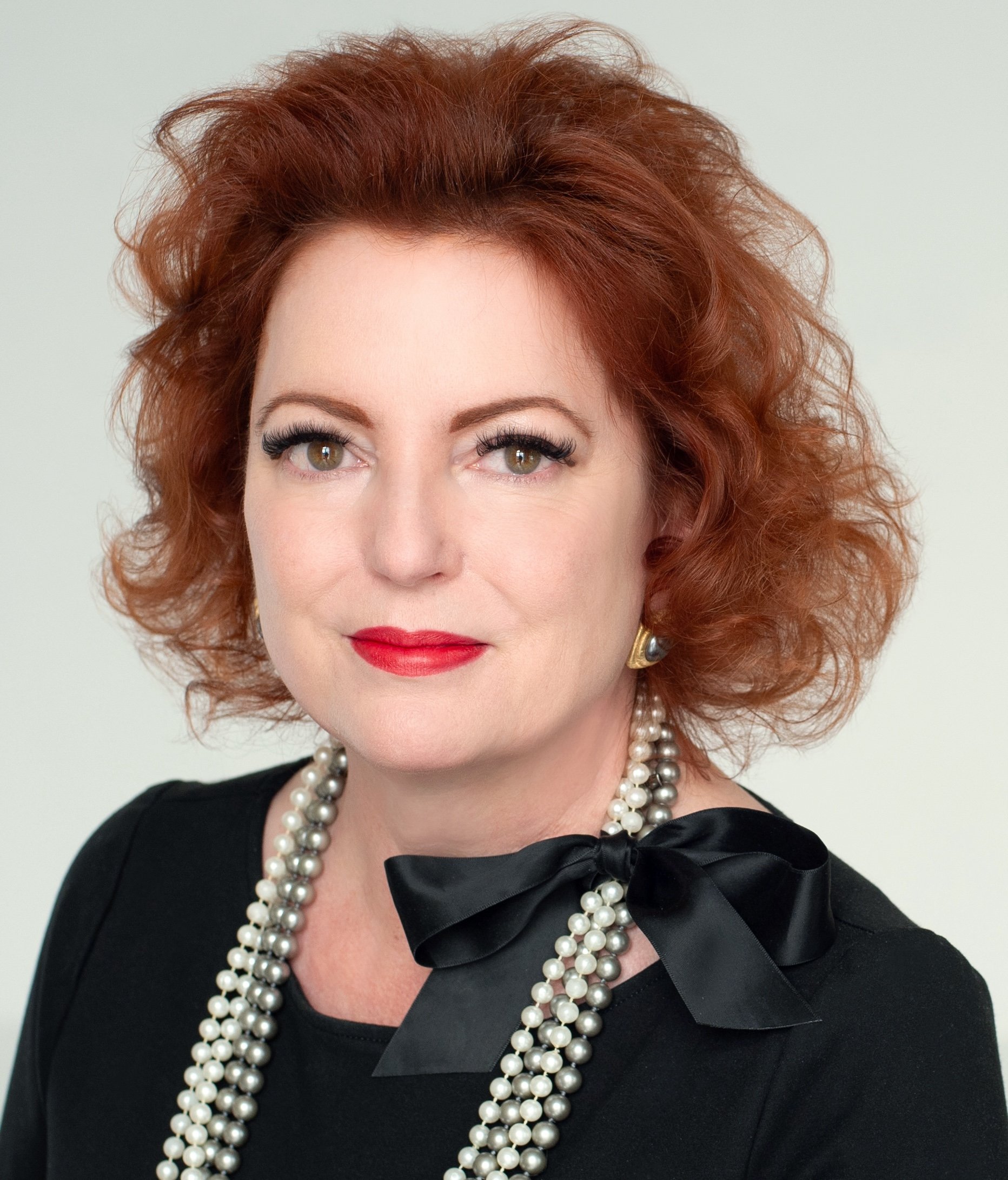 Close-up of a woman with red curly hair, wearing a black dress with a bow and a layered pearl necklace.