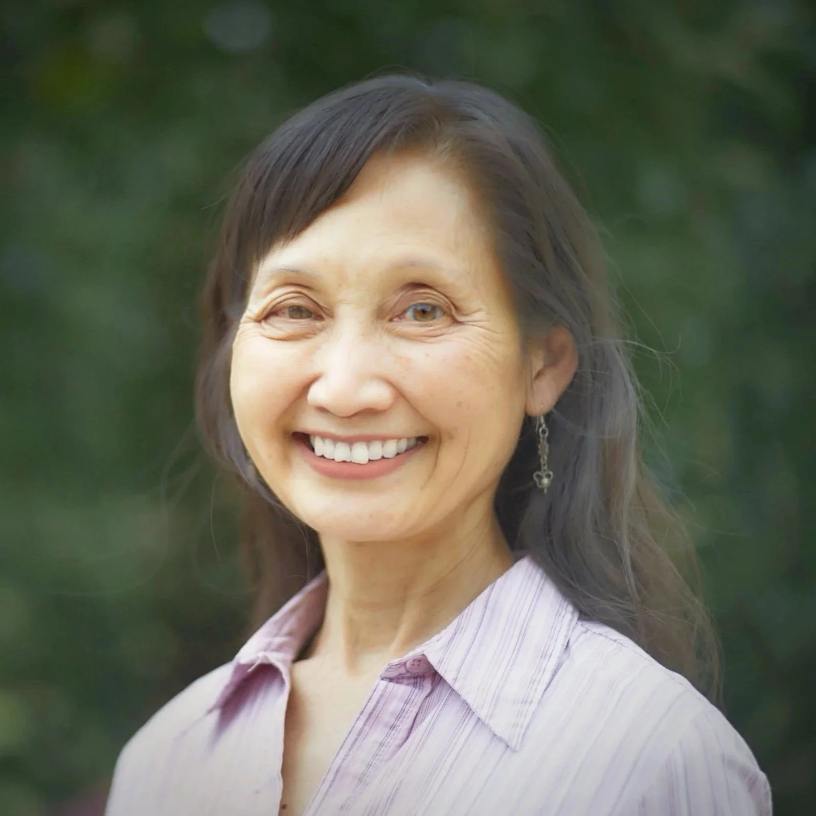 A smiling woman with long dark hair, wearing a light striped shirt, standing outdoors with a blurred green background.