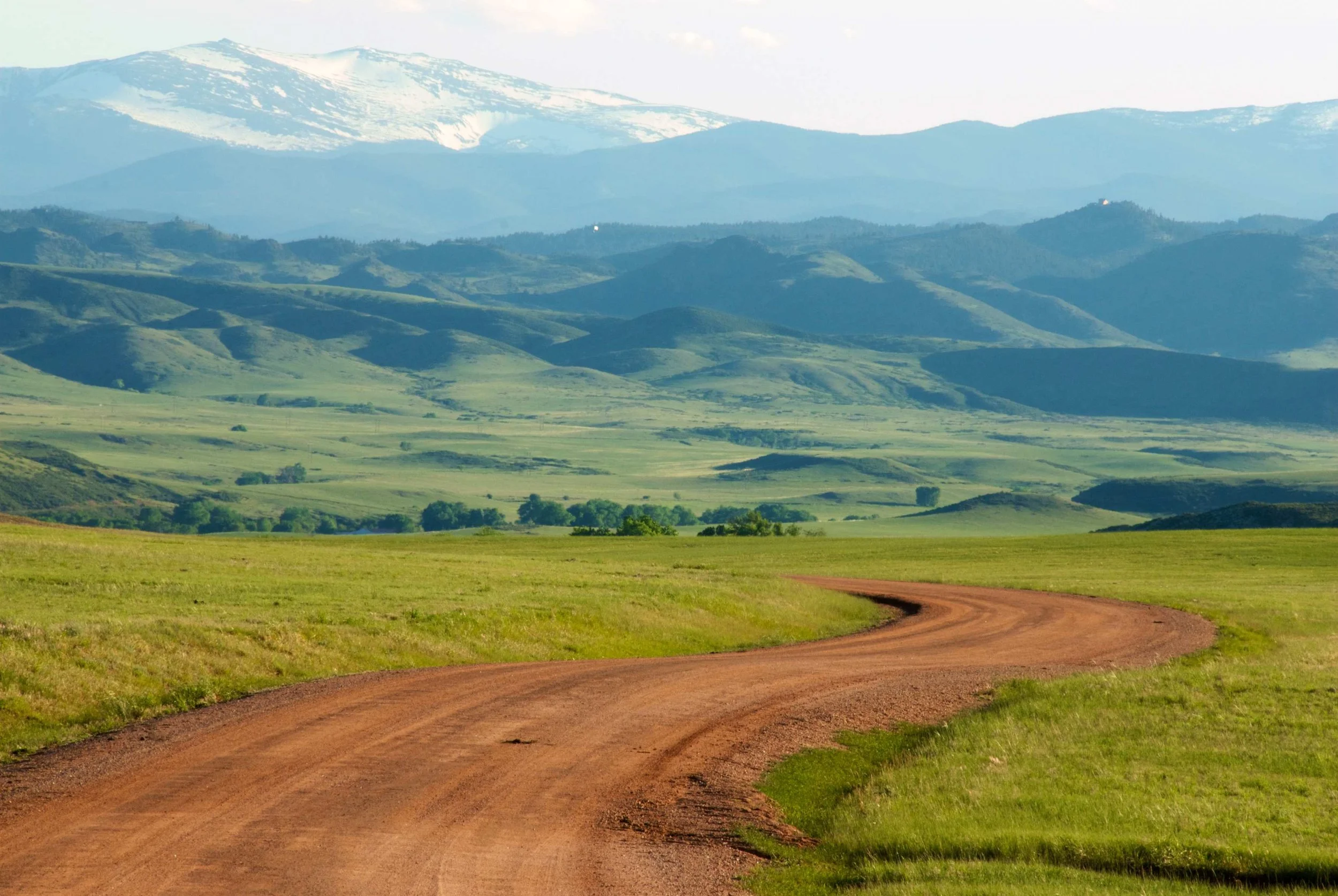 Rural landscape with a dirt road winding through green fields and rolling hills, snow-capped mountains in the background under a clear sky.