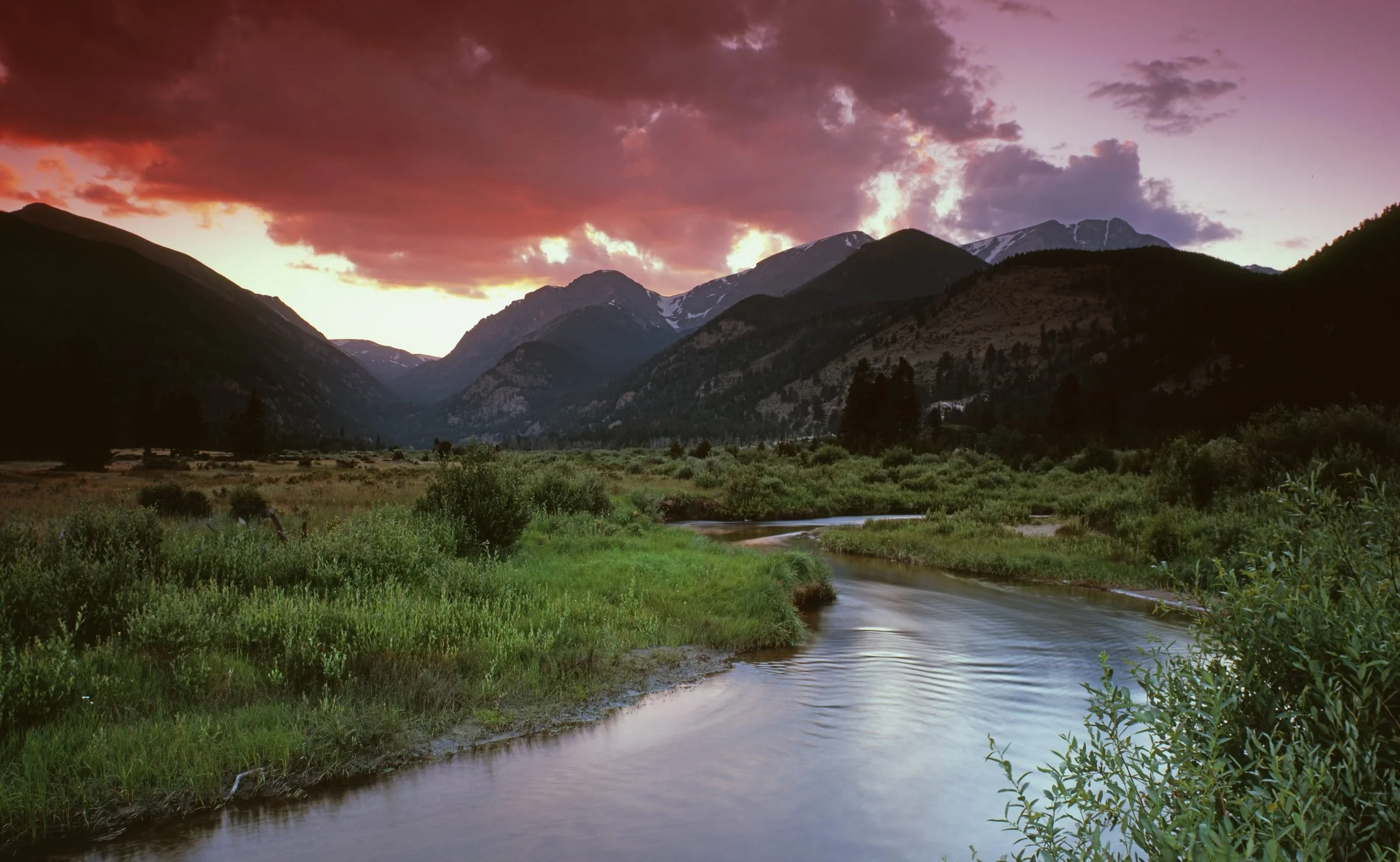 A scenic landscape of mountain ranges during sunset with dark clouds, lush green grasses and a flowing river in the foreground.