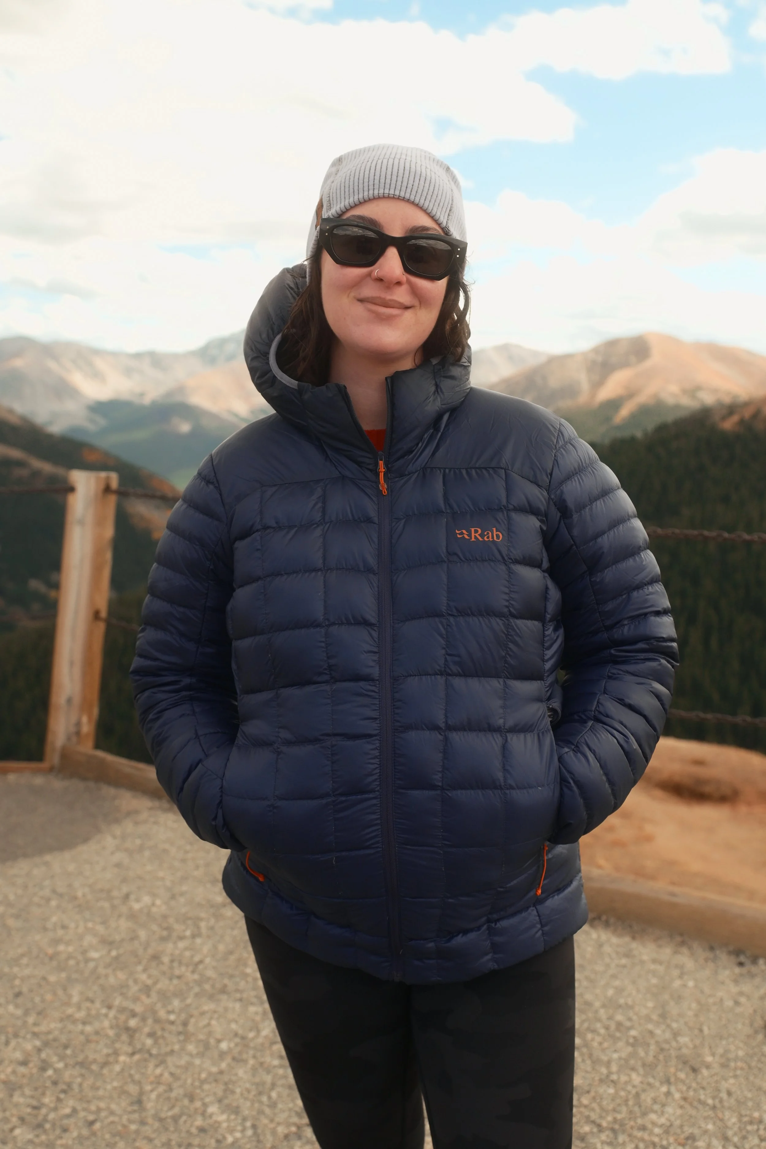 A woman wearing a gray beanie, sunglasses, and a navy puffer jacket standing outdoors with mountains in the background.