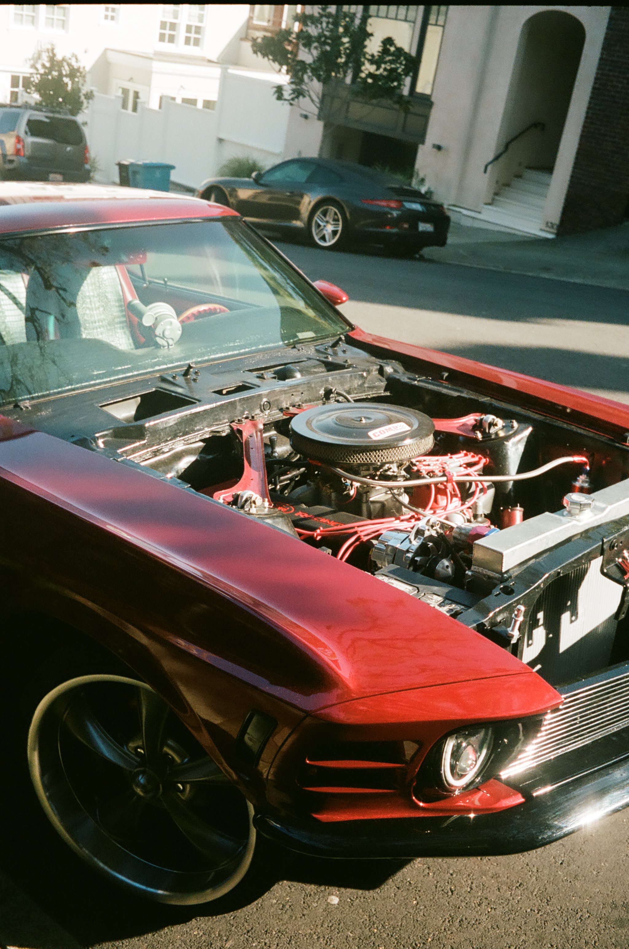 Red classic Ford Mustang with the hood open, exposing the engine, parked on a residential street with other cars and houses in the background.