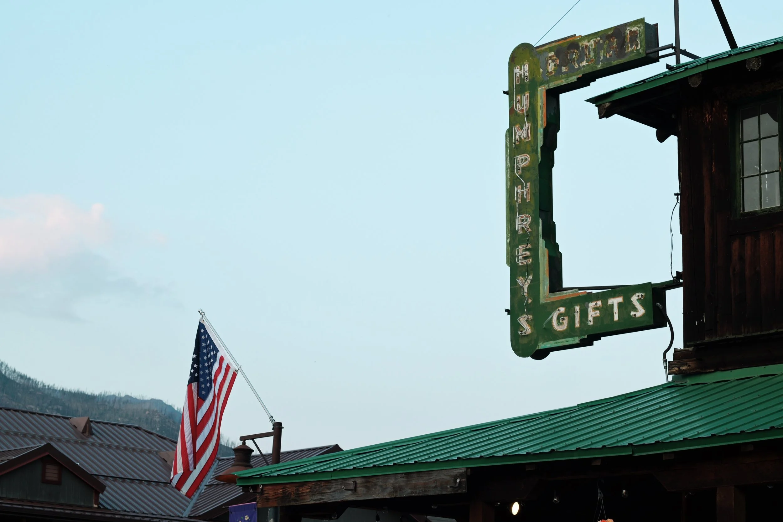 An outdoor scene with a vintage green neon sign for 'Gifts & More' attached to a wooden building with a green metal roof, and an American flag on a pole in the background.