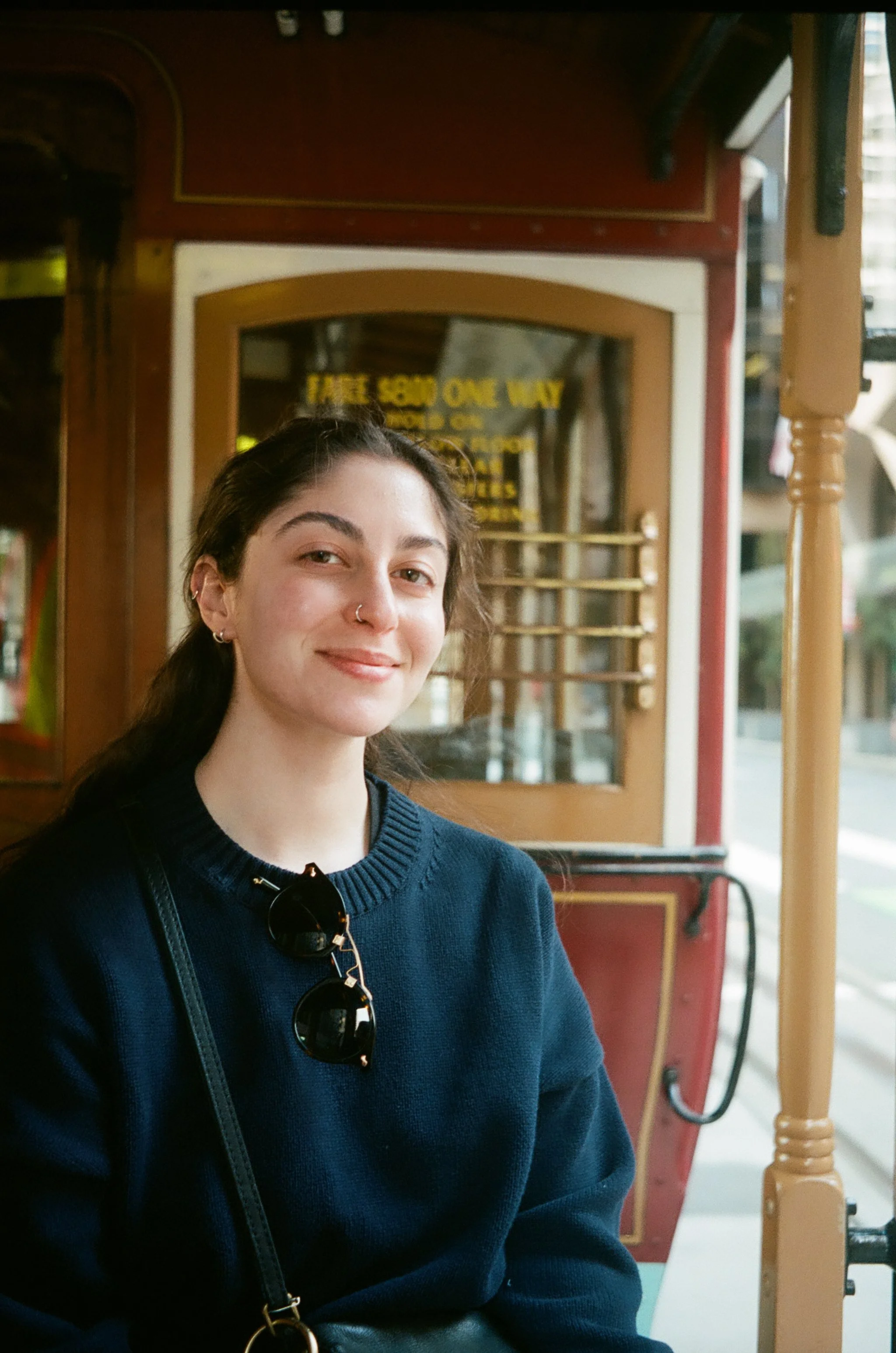 A young woman with dark hair, wearing a navy blue sweater, has sunglasses hanging from her sweater and a nose ring. She is sitting inside a vintage-style trolley, smiling at the camera.
