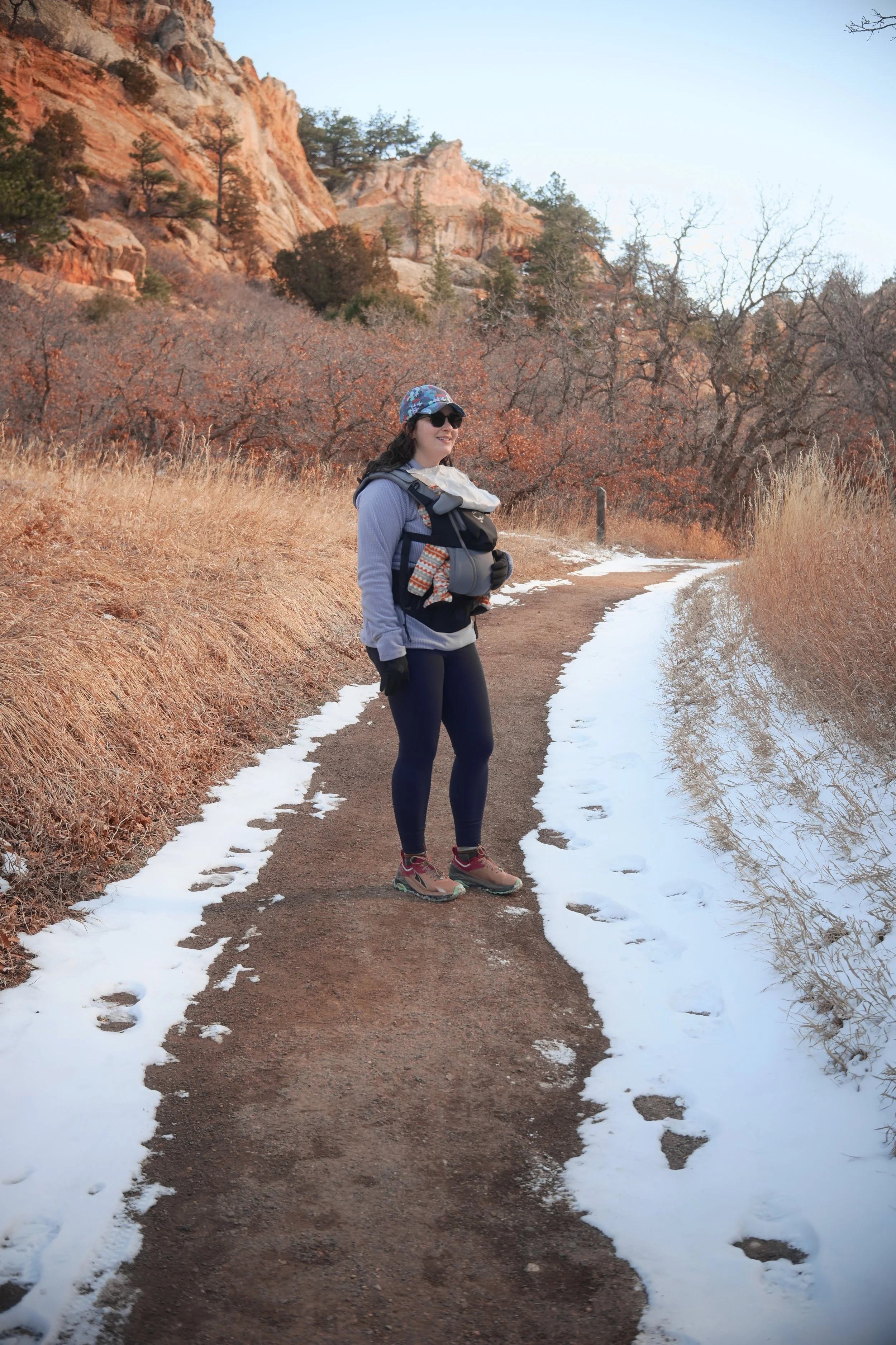 A woman hiking on a trail with snow on the sides, surrounded by dry grasses and trees in a mountainous area.