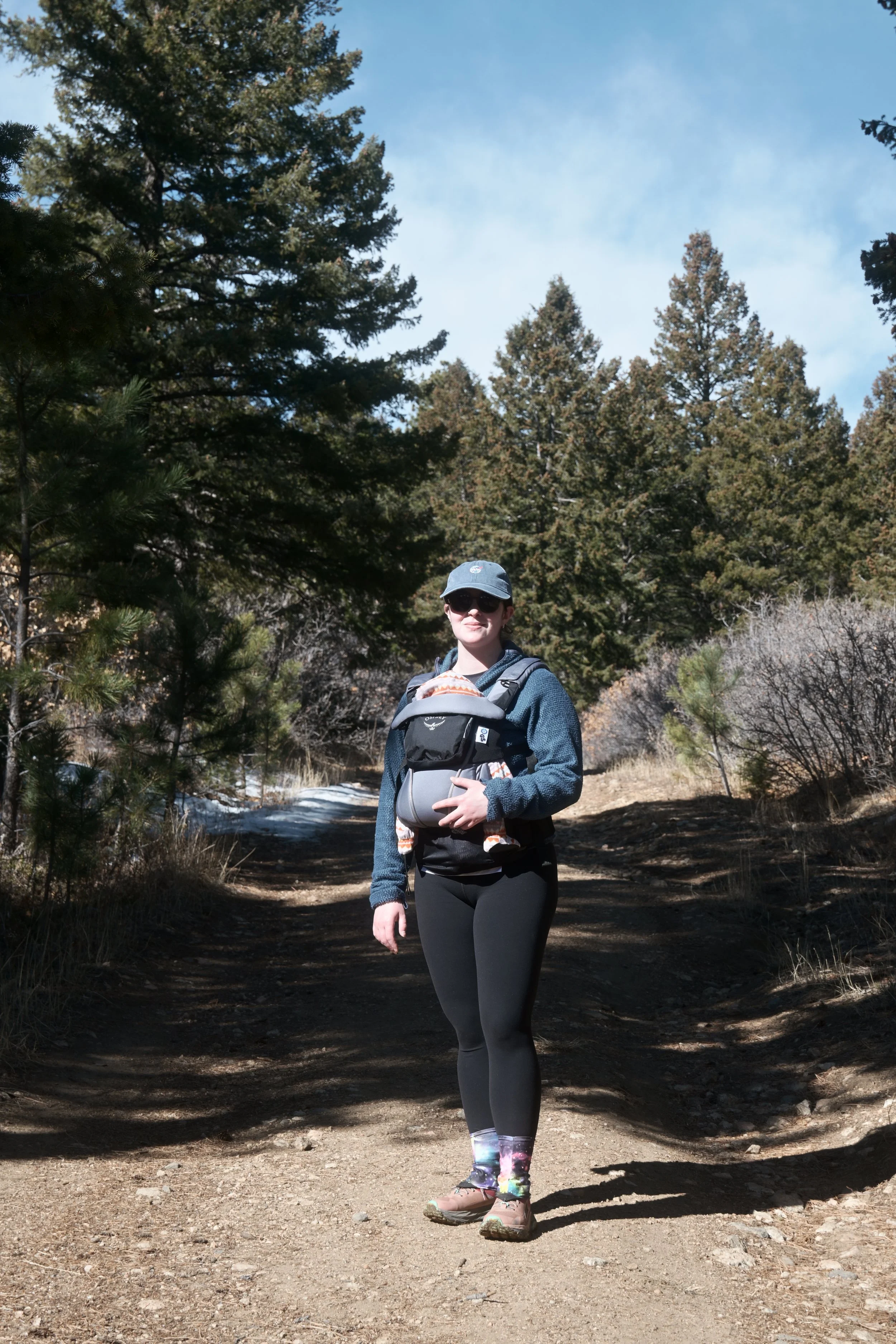 Woman hiking on a trail in the woods, wearing sunglasses, a hat, a backpack, leggings, and hiking boots, with pine trees and a partly cloudy sky in the background.