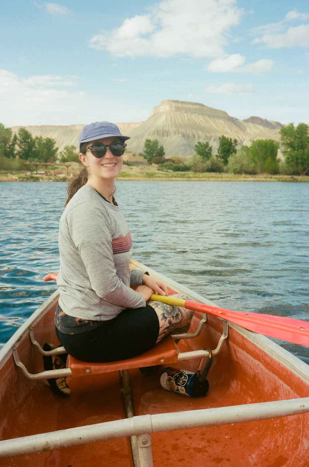 A woman with sunglasses and a hat sitting in a canoe on a lake, holding a paddle, with a backdrop of a mountain and a partly cloudy sky.