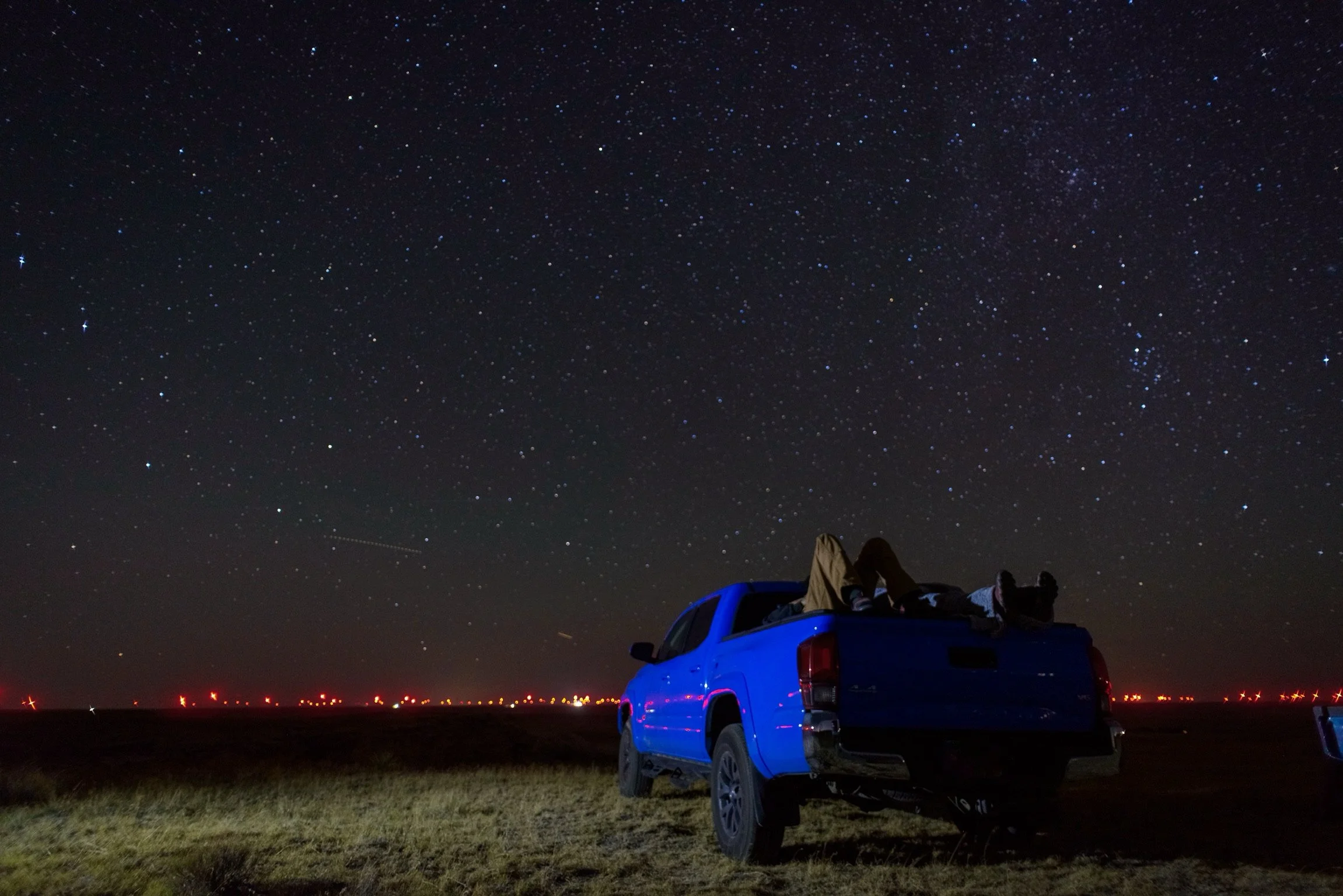 A person lying in the back of a blue pickup truck under a starry night sky with visible constellations and a faint trail from a passing airplane.