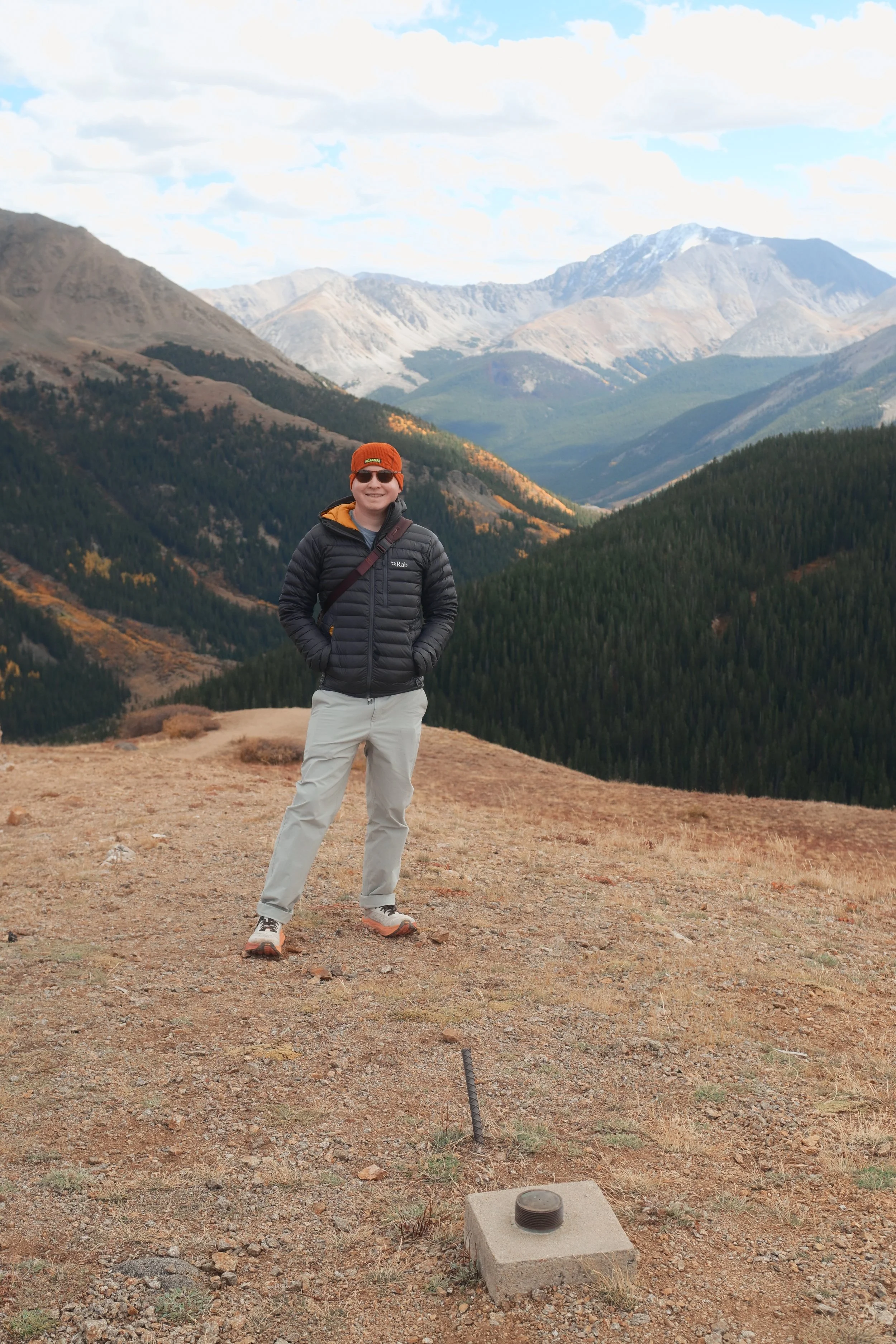 Man standing on mountain trail with mountains and forest in background, wearing orange beanie, sunglasses, black puffy jacket, and beige pants.