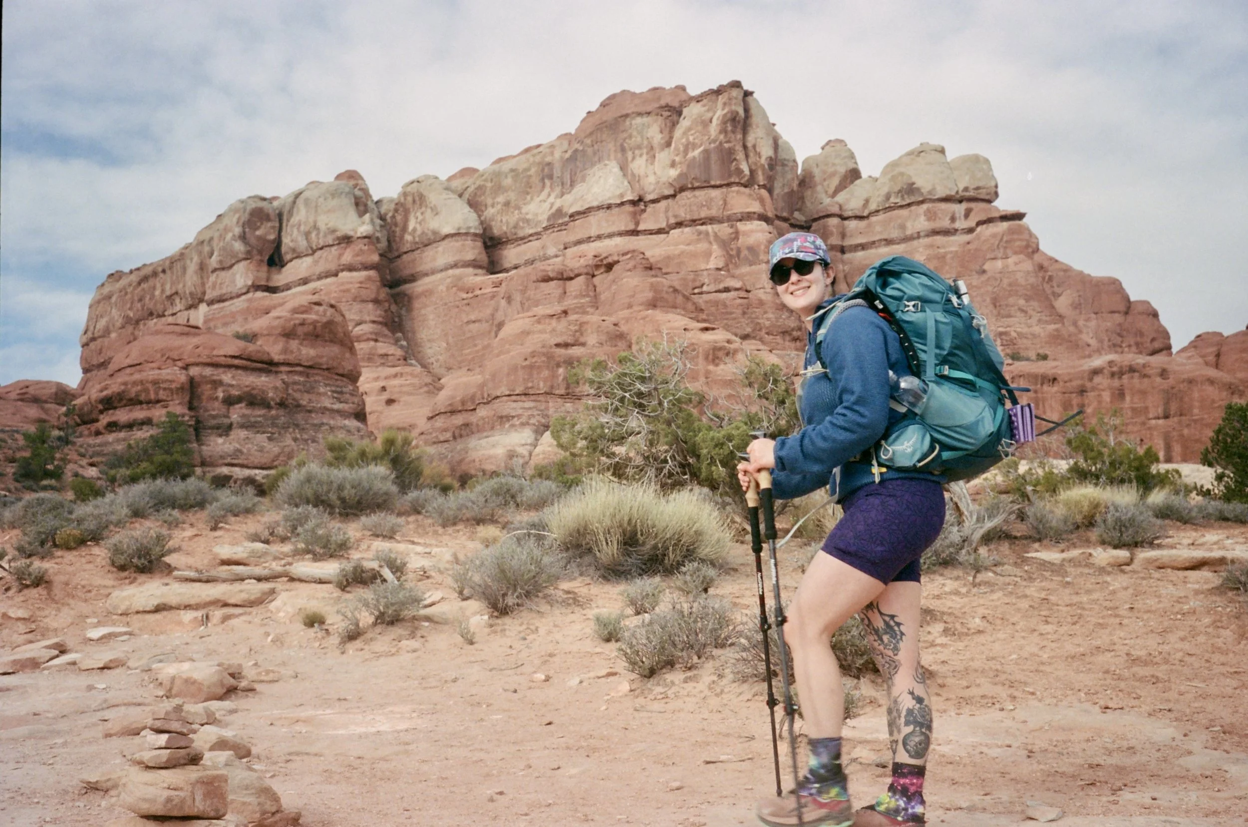 smiling woman hiking with a large backpack and trekking poles in a desert landscape with red rock formations and sparse vegetation