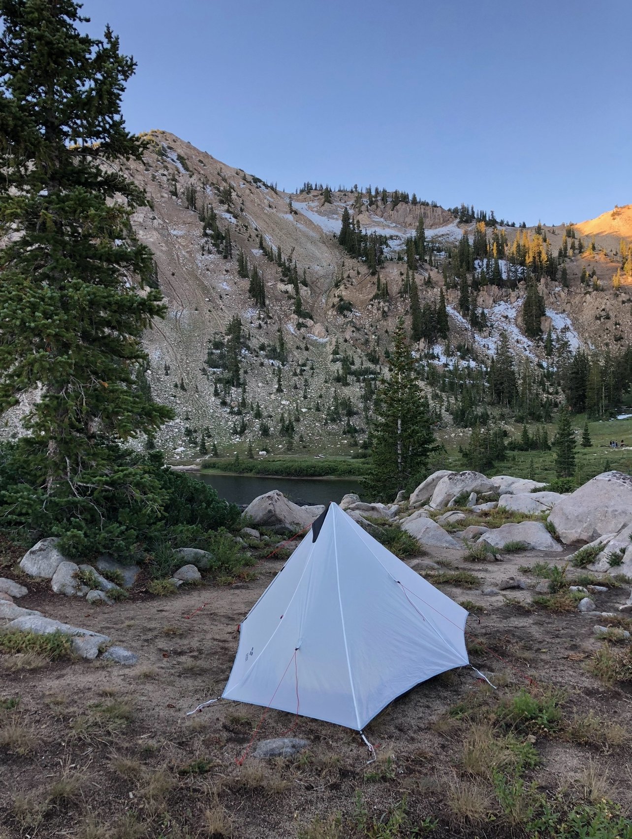 A white tent set up on a grassy patch with rocks, near a lake, surrounded by tall pine trees, mountains with patches of snow, under a clear blue sky.