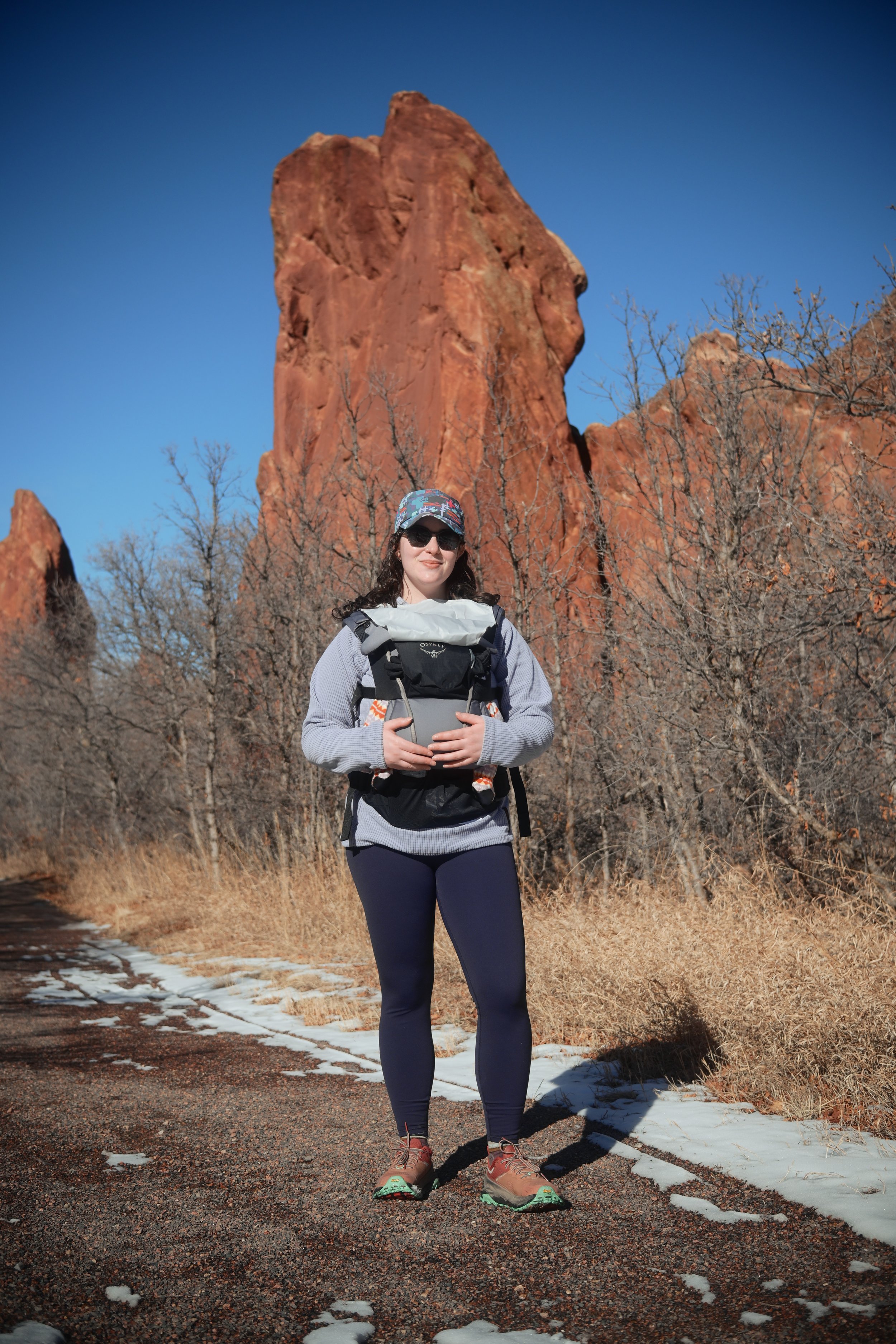 A woman standing on a trail with red rock formations and leafless trees in the background, wearing hiking gear including a backpack, hat, sunglasses, gray sweater, dark leggings, and hiking boots on a sunny day.