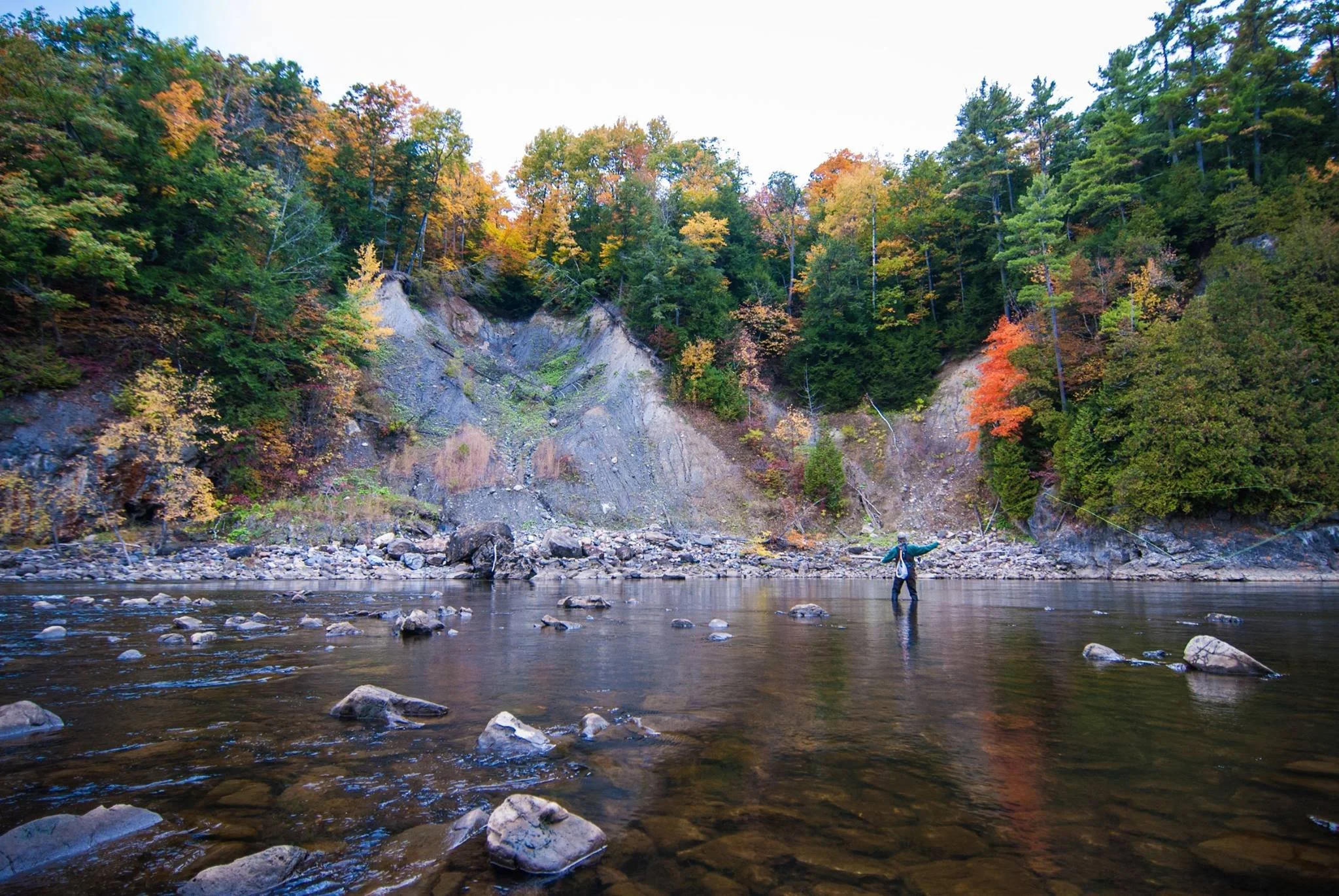 A person fishing in a river with rocky banks, surrounded by trees with colorful autumn foliage.
