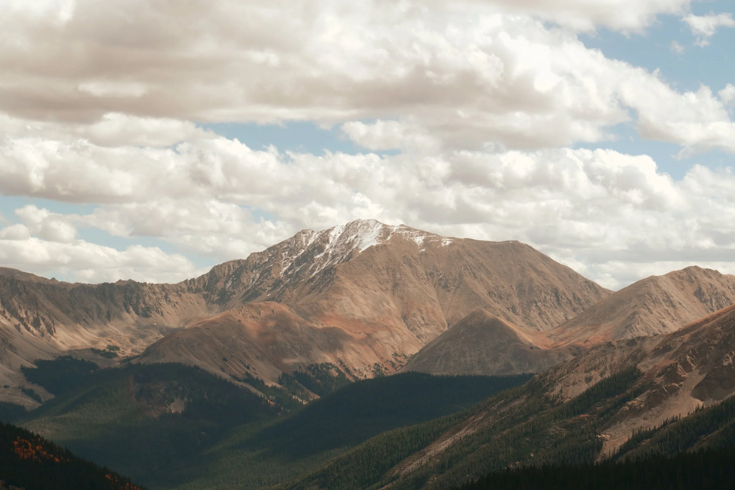 Mountain landscape Independence Pass with snow-topped peak, rugged brown slopes, and green forests in the foreground under a partly cloudy sky.