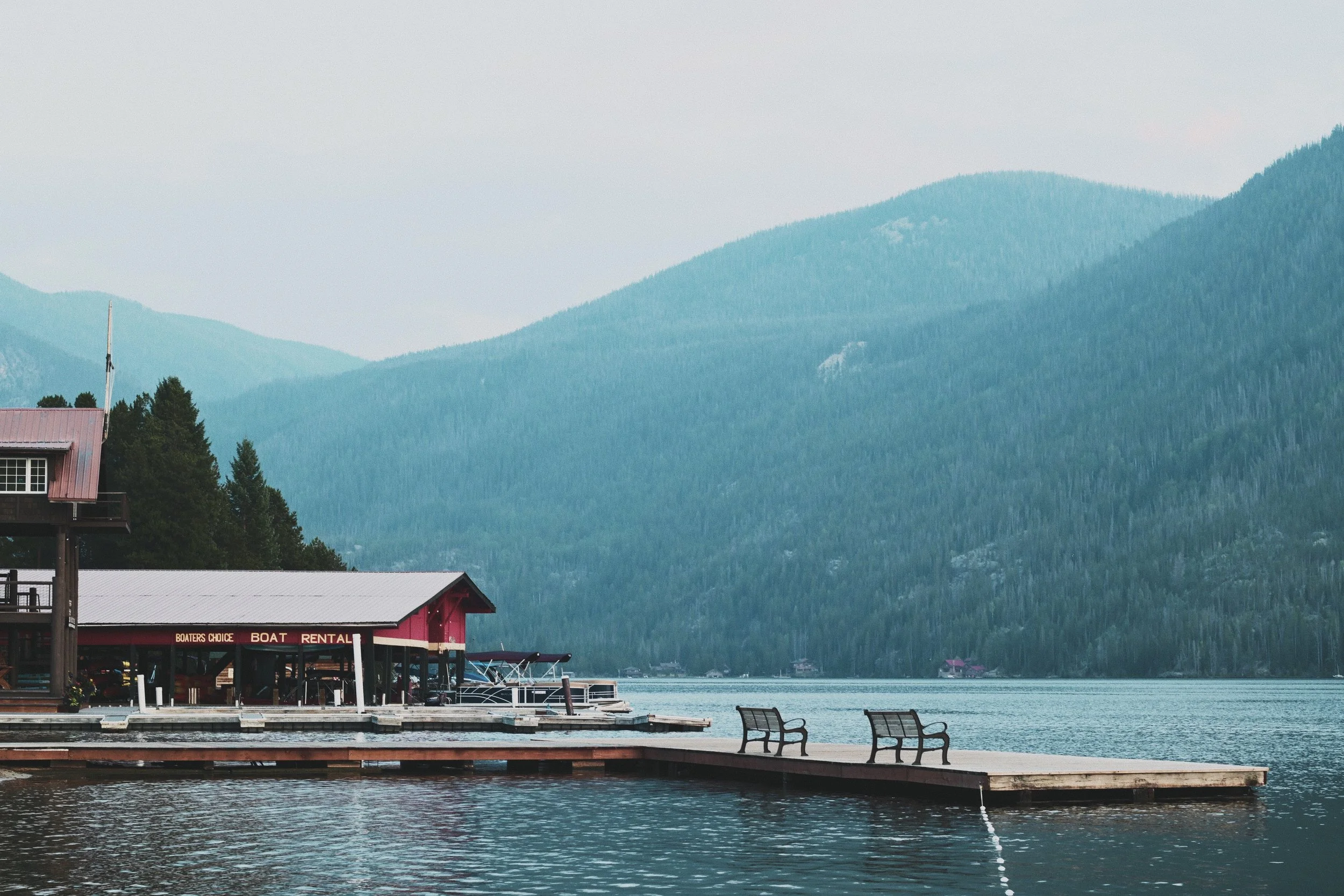 A lakeside dock in Grand Lake Colorado with two empty benches in front, a boat rental shop with boats inside, and lush green mountains in the background.