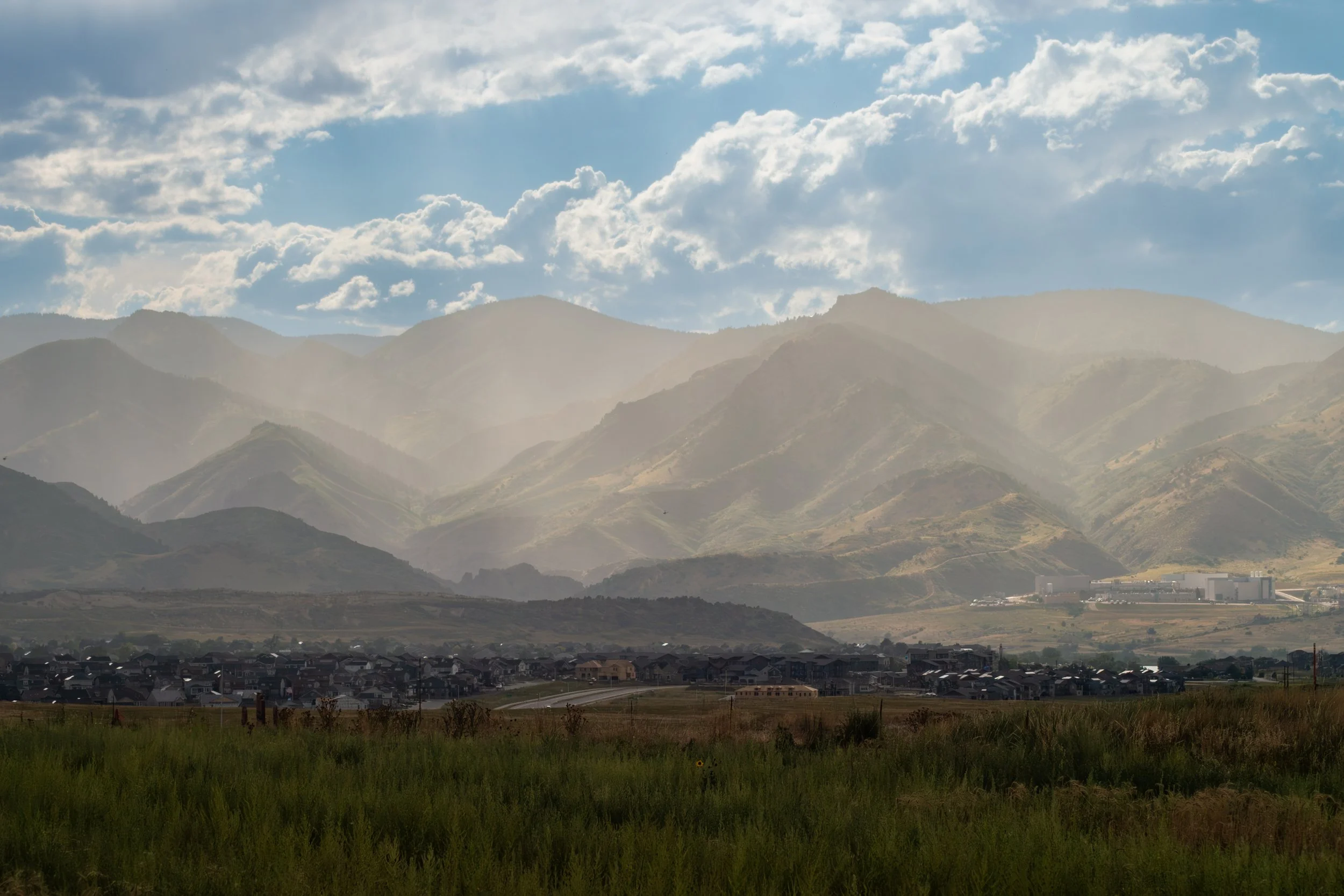 A landscape with rolling green hills in the foreground, a small town or city nestled at the base, and mountain ranges in the background under a partly cloudy sky with sunlight streaming through.