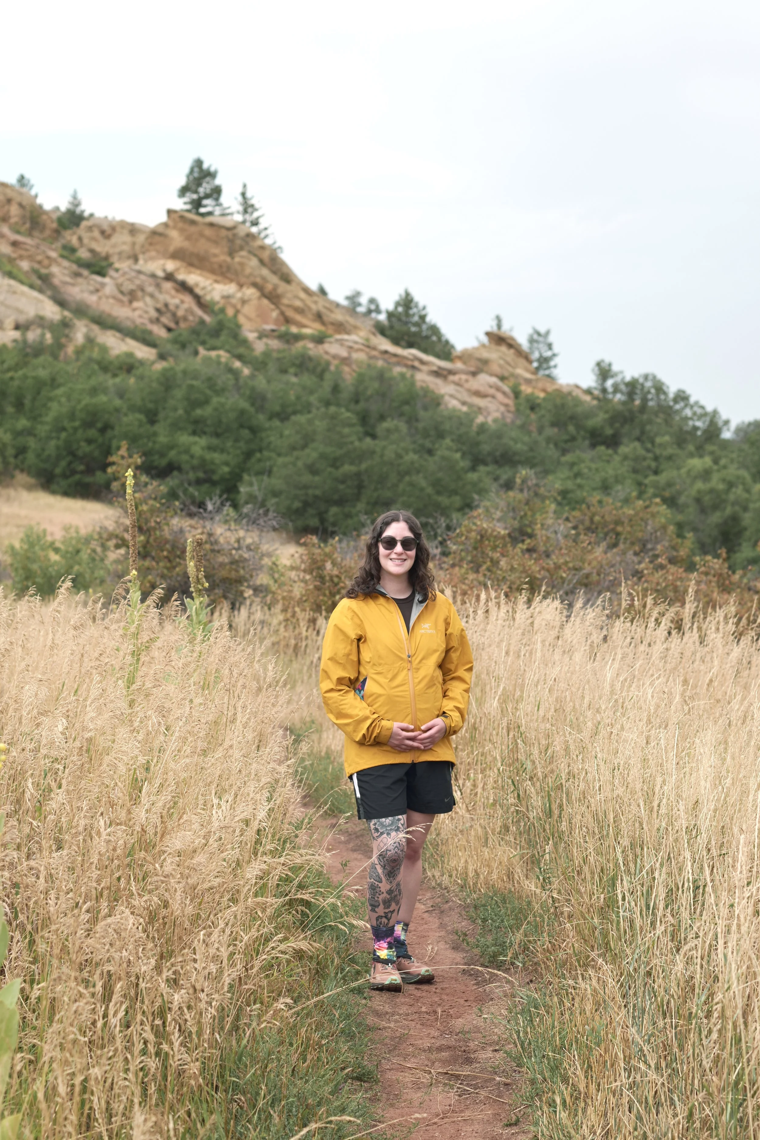 A woman wearing sunglasses, a yellow jacket, black shorts, colorful socks, and hiking boots standing on a dirt trail in a grassy field with rocky hills and trees in the background.