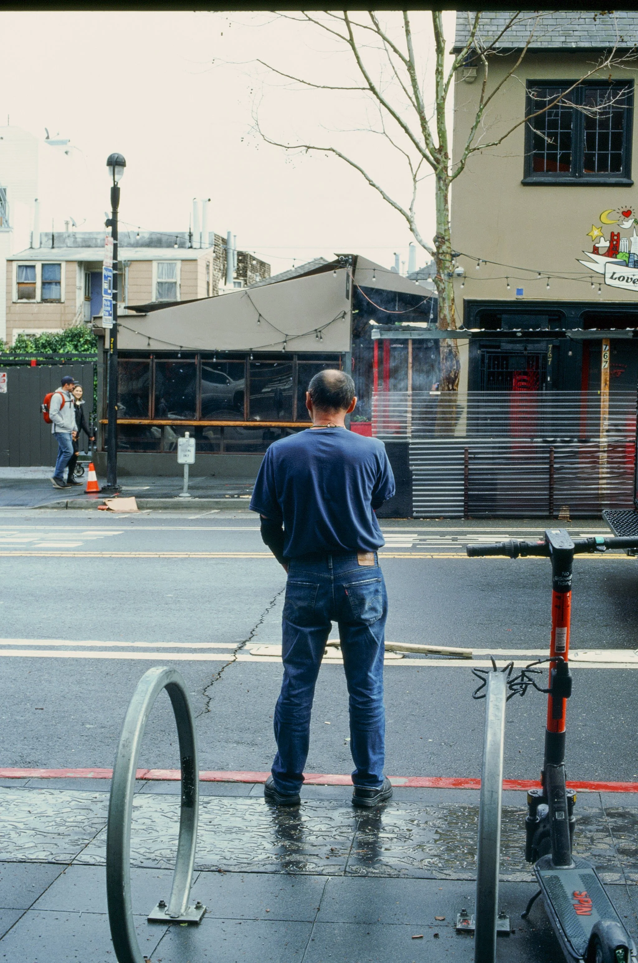 A man standing on a city sidewalk, facing away, observing a street scene with pedestrians, buildings, and a tree, during overcast weather.