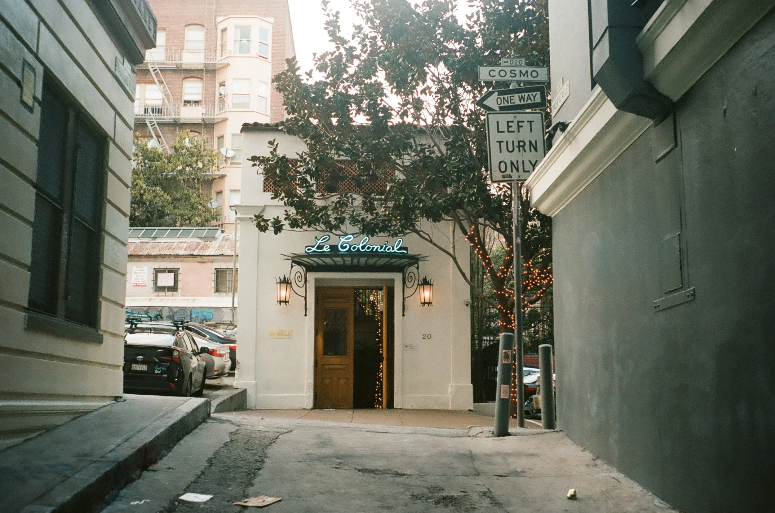 An alleyway with a white building named Le Colonial, decorated with string lights, and parking cars on the side. Street signs indicate directions for Cosmo, one way, and left turn only.
