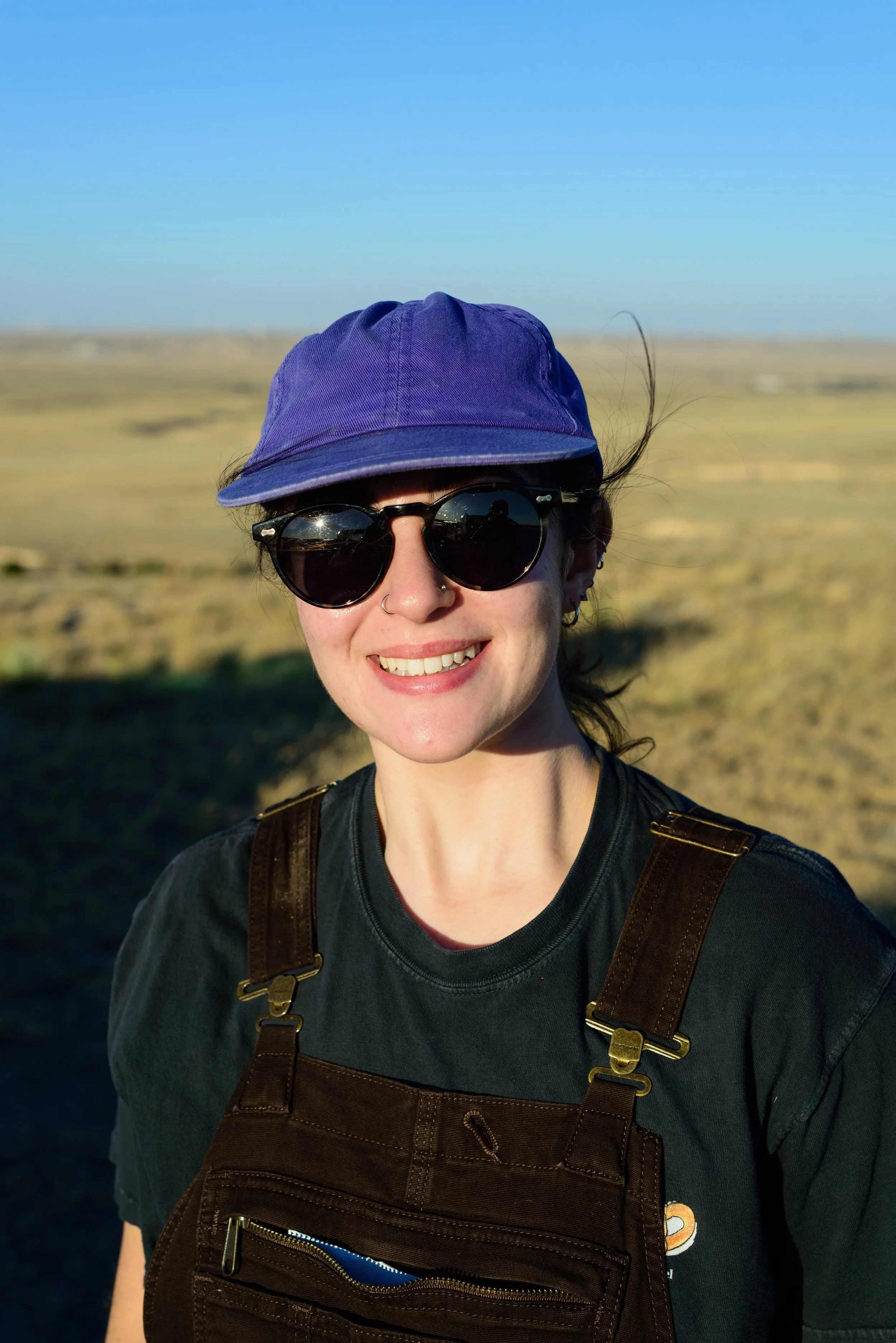 Woman wearing sunglasses, purple cap, and brown overalls outdoors in a vast open landscape with clear blue sky.