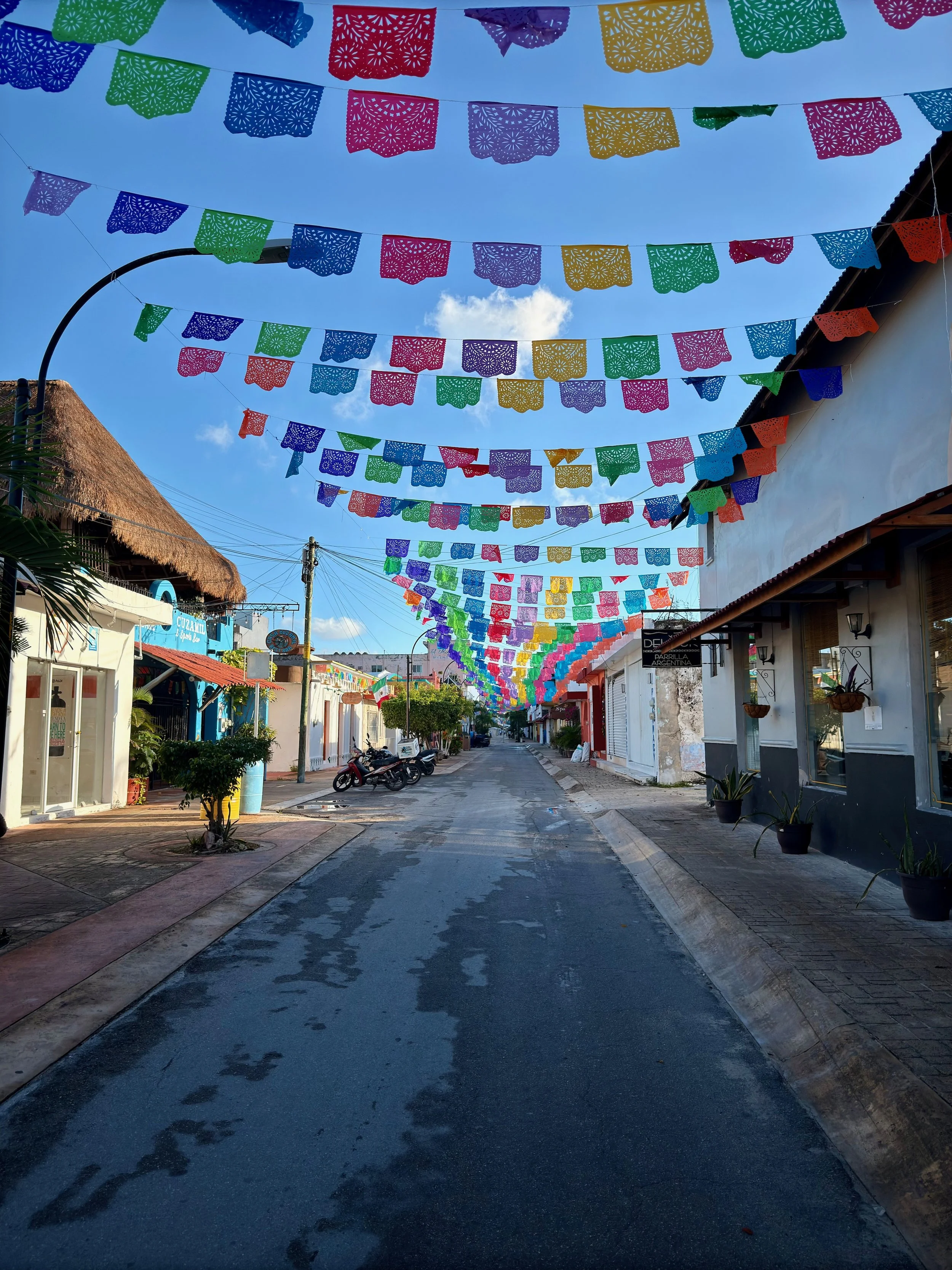 Colorful papel picado banners hanging across a street on a sunny day, with buildings and motorcycles on the sides.