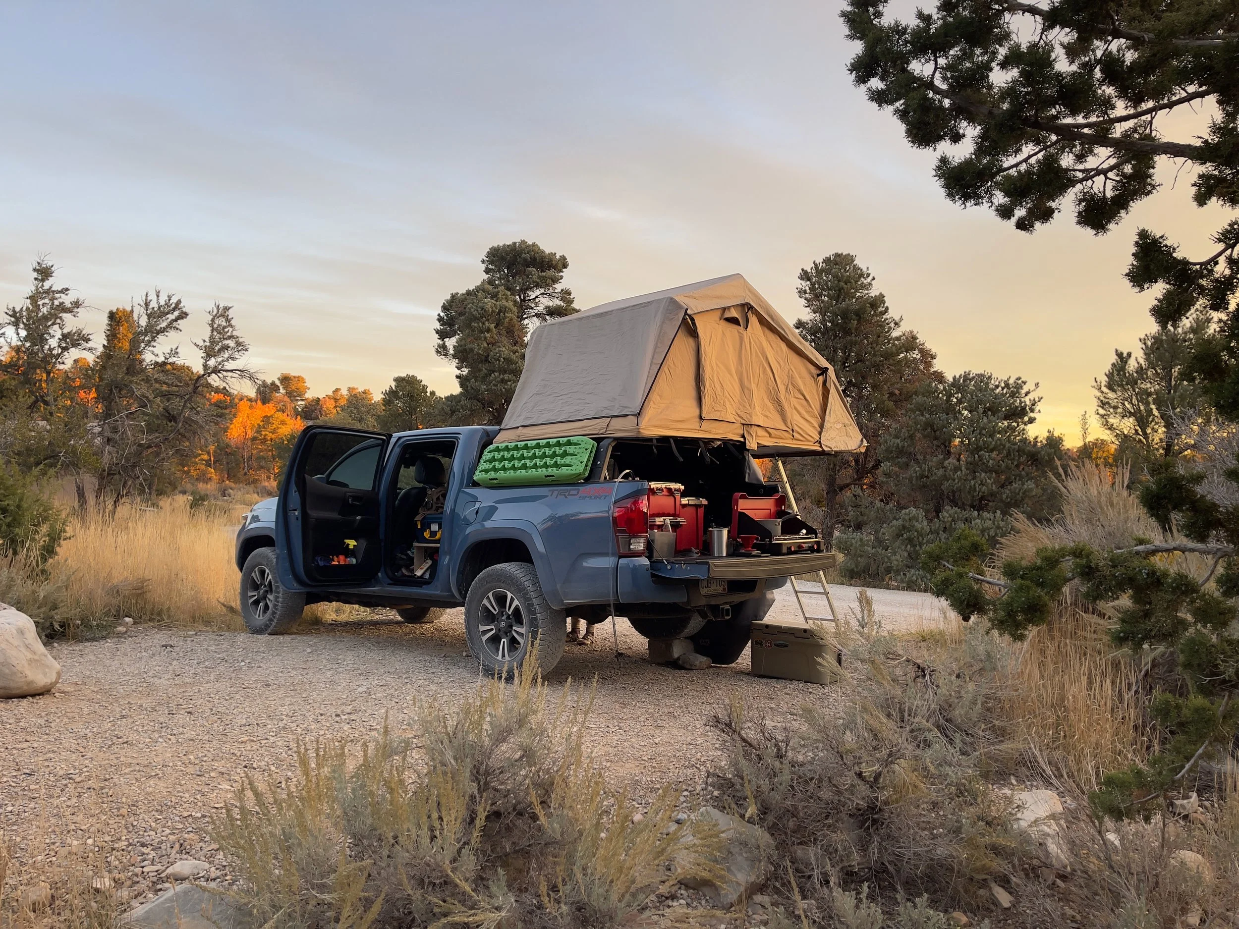 A blue pickup truck with an open door and a roof-top tent parked on a dirt trail in a desert landscape with trees and shrubs, during sunset.