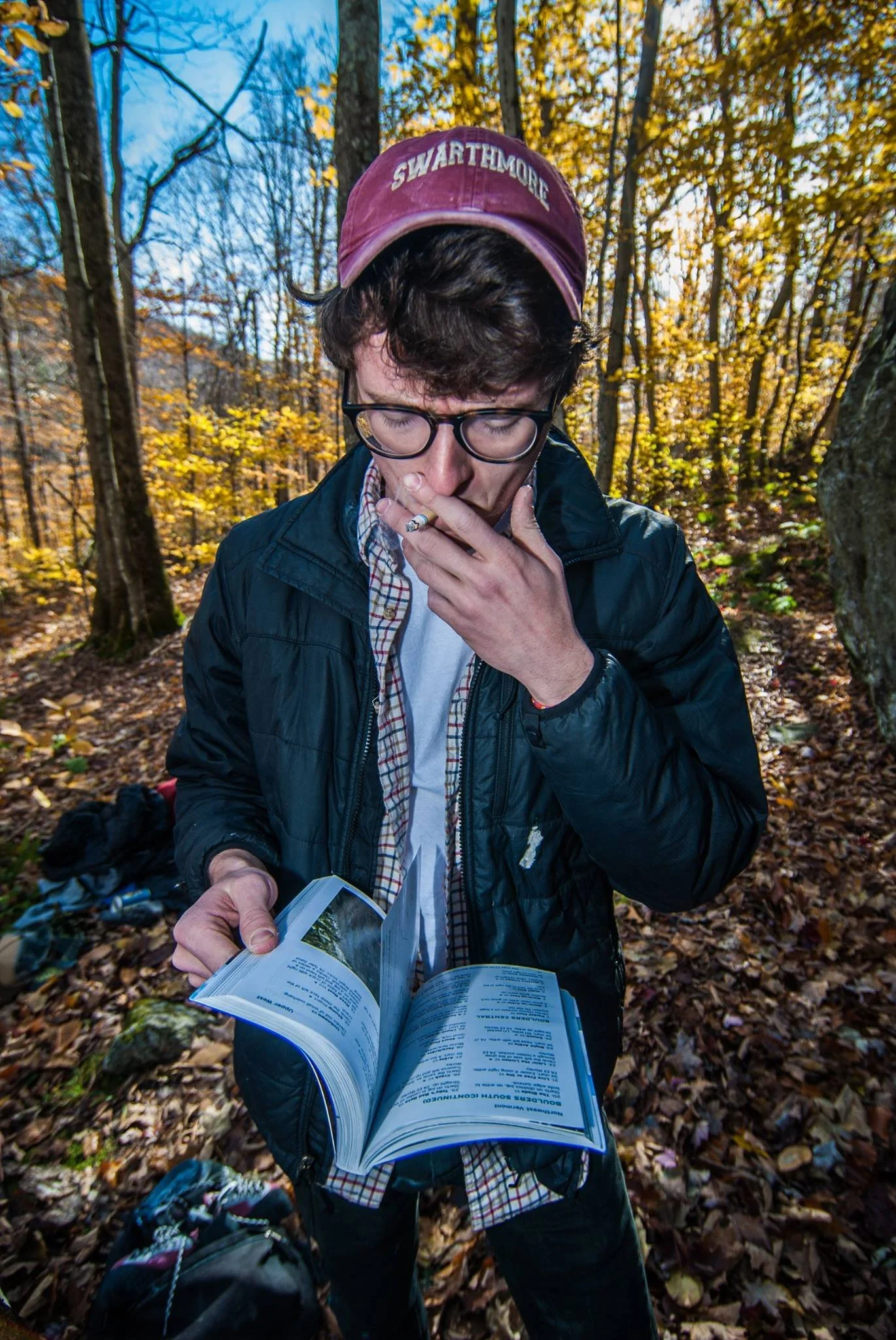 A young man with glasses wearing a maroon cap, black jacket, and a plaid shirt, holding an open guidebook and smoking a cigarette in a forest during fall with colorful leaves and trees in the background.