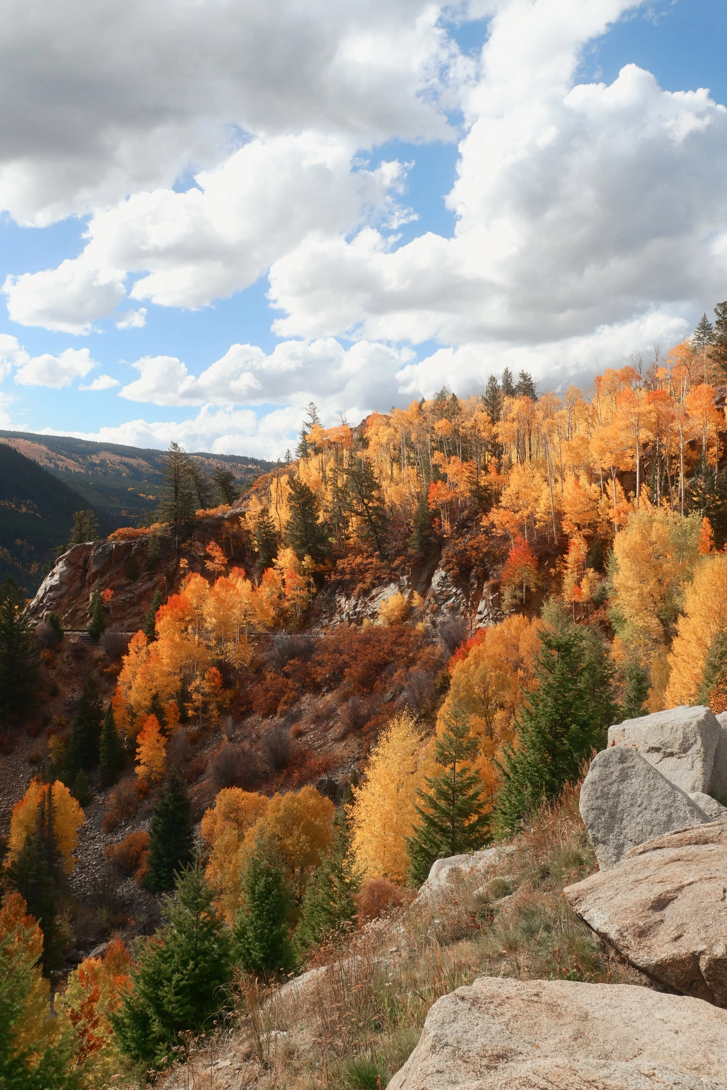 A scenic mountain landscape with colorful fall foliage, rocky terrain, and a partly cloudy sky.