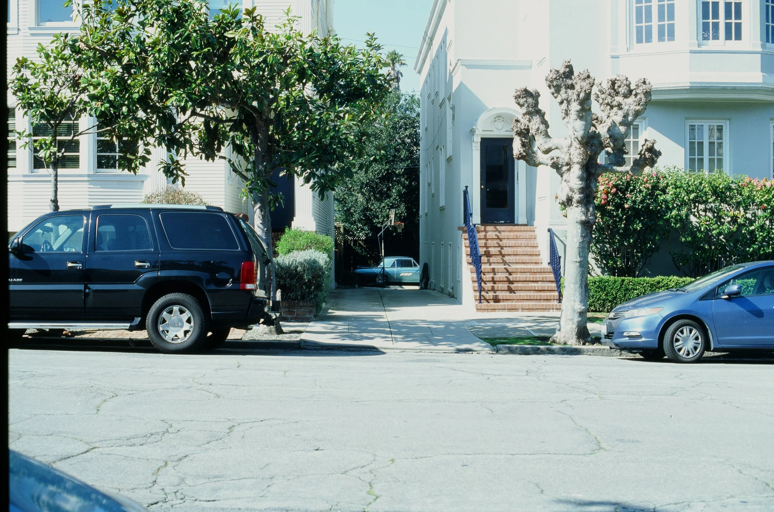 Front view of a white residential building with brick stairs, two trees, and parked cars on the street.