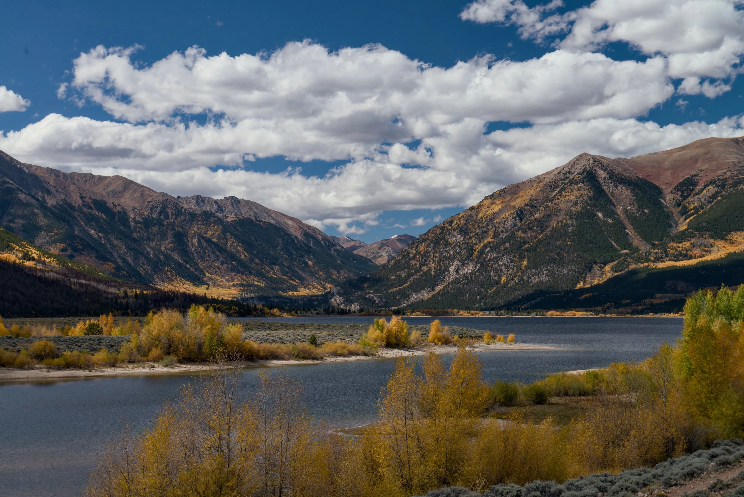 Scenic view of a mountain landscape with Twin Lakes Colorado in the foreground and mountains in the background, under a partly cloudy sky, with trees in fall colors along the shore.